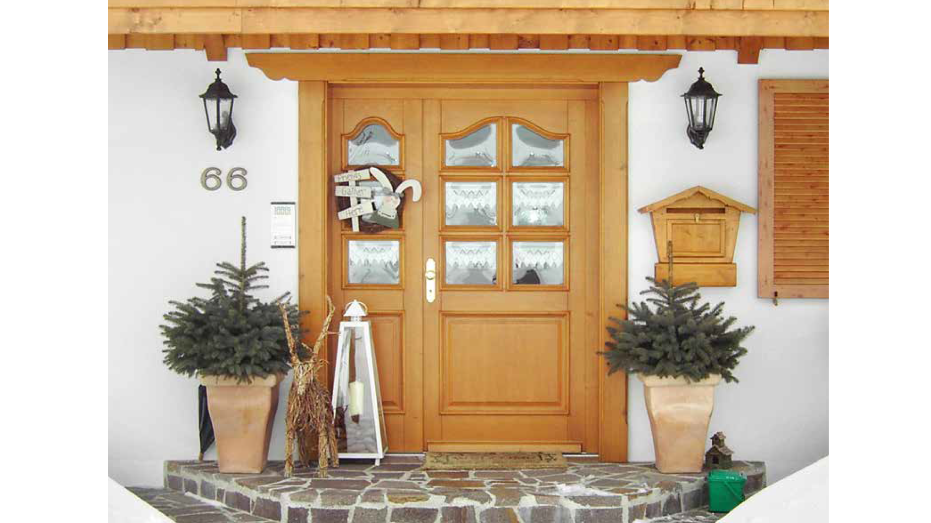 Holztür mit Glaseinsätzen, seitlich und darüber zwei Wandlampen, zwei Tannenbäume in Tontöpfen, Weihnachtsschmuck, Hausnummer 66, Briefkasten, Willkommensschild, Türmatte, vor der Tür ein gemauerter Vorgarten