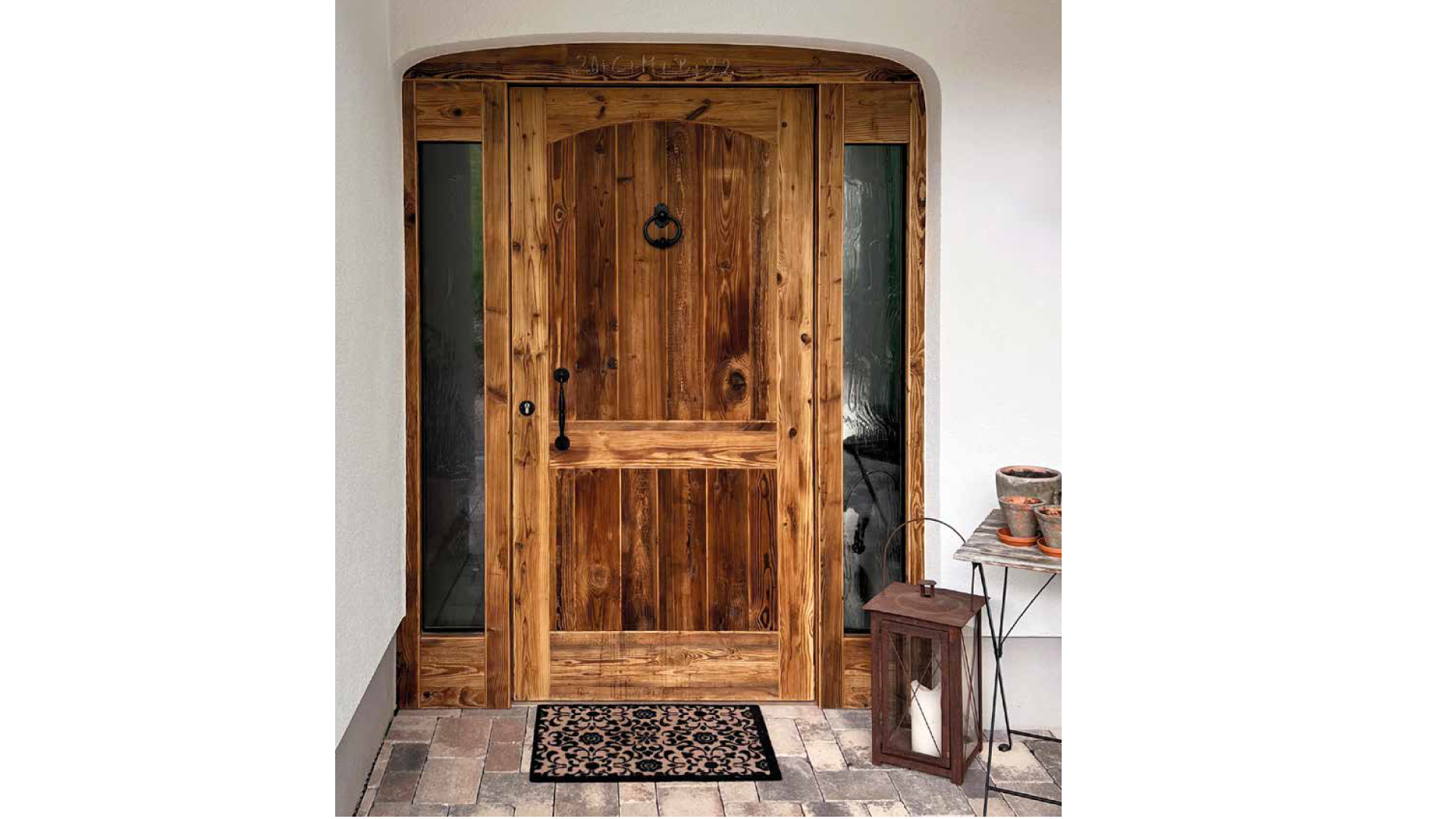 Holztür mit schwarzen Beschlägen vor weißer Wand, umgeben von Seitengläsern, mit einem begrünten Innenhof im Hintergrund, auf terrakottafarbenem Pflaster, mit einem schwarzen und beige gemusterten Fußmatte davor, rechtsseitig eine kleine Holzkiste mi