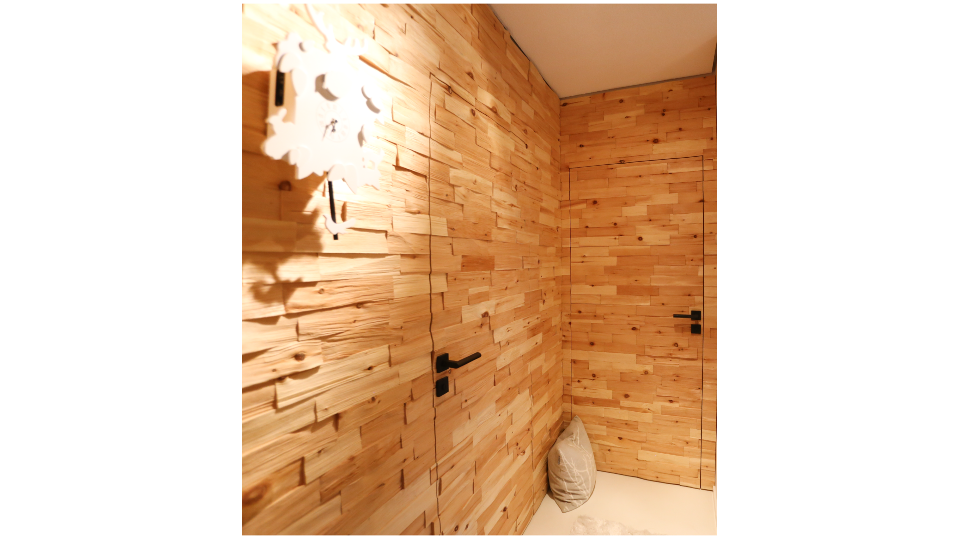 Ein Flur mit Wänden aus Holzvertäfelung, zwei schwarze Türgriffe, eine Wandlampe und ein Sitzkissen auf dem Boden in der Ecke.