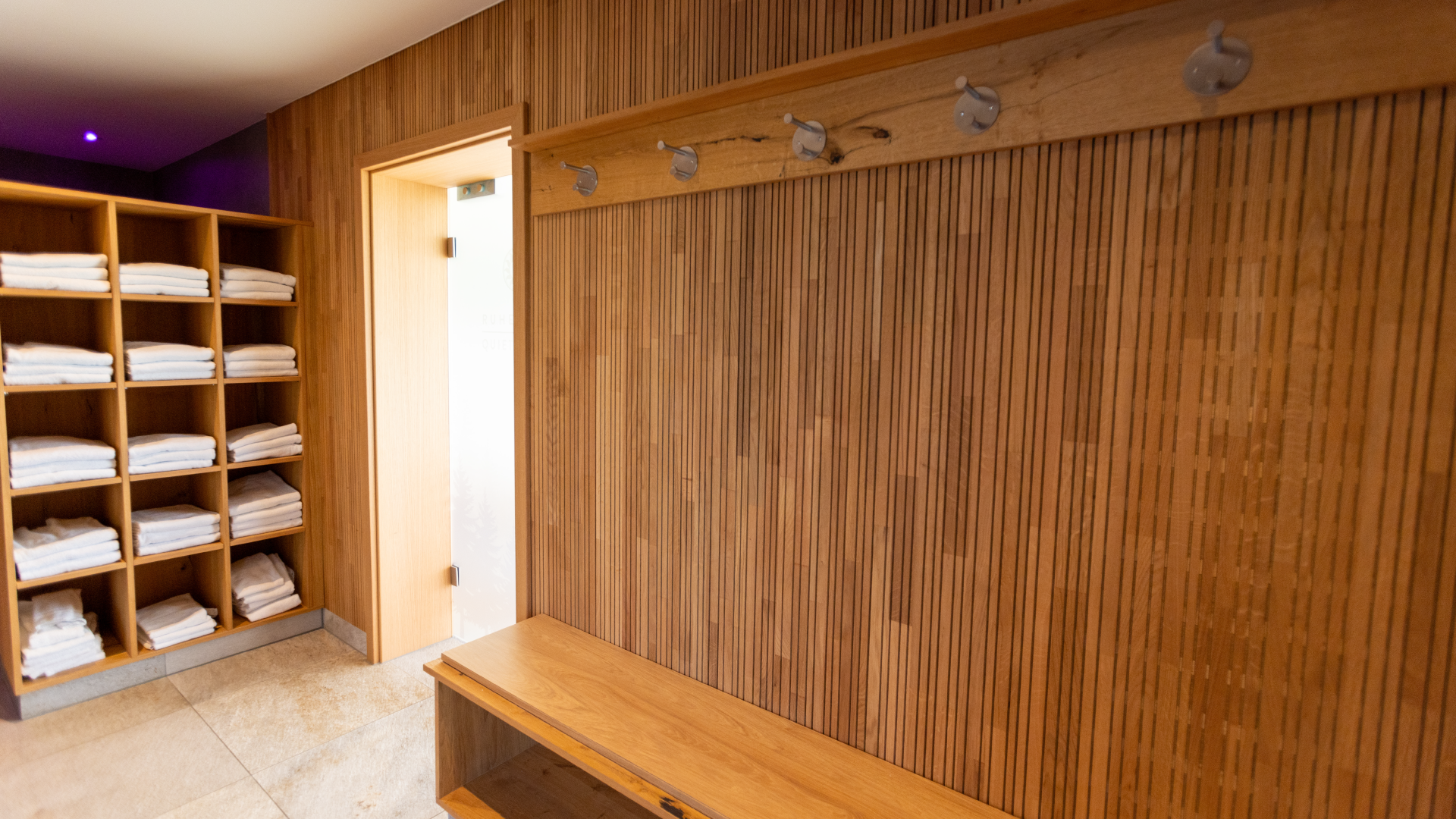 Holzwand mit Hakenleiste und Ablage, daneben offene Schränke mit schwarz-weißen Handtüchern in einer Sauna oder Spa-Umgebung.
