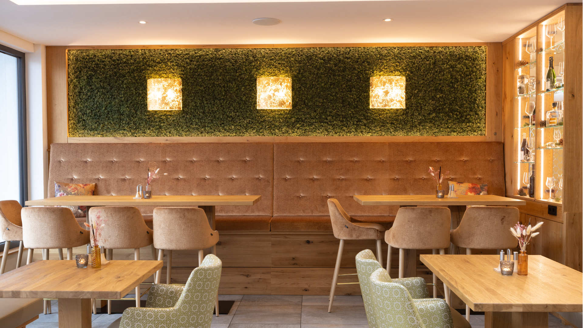 Gemütliches Restaurant mit Holzdekor, gepolsterten Sitzbänken, einzelnen Stühlen, Tischen mit kleinen Dekorationen und einer grünen Wand mit drei quadratischen Lichtornamenten.