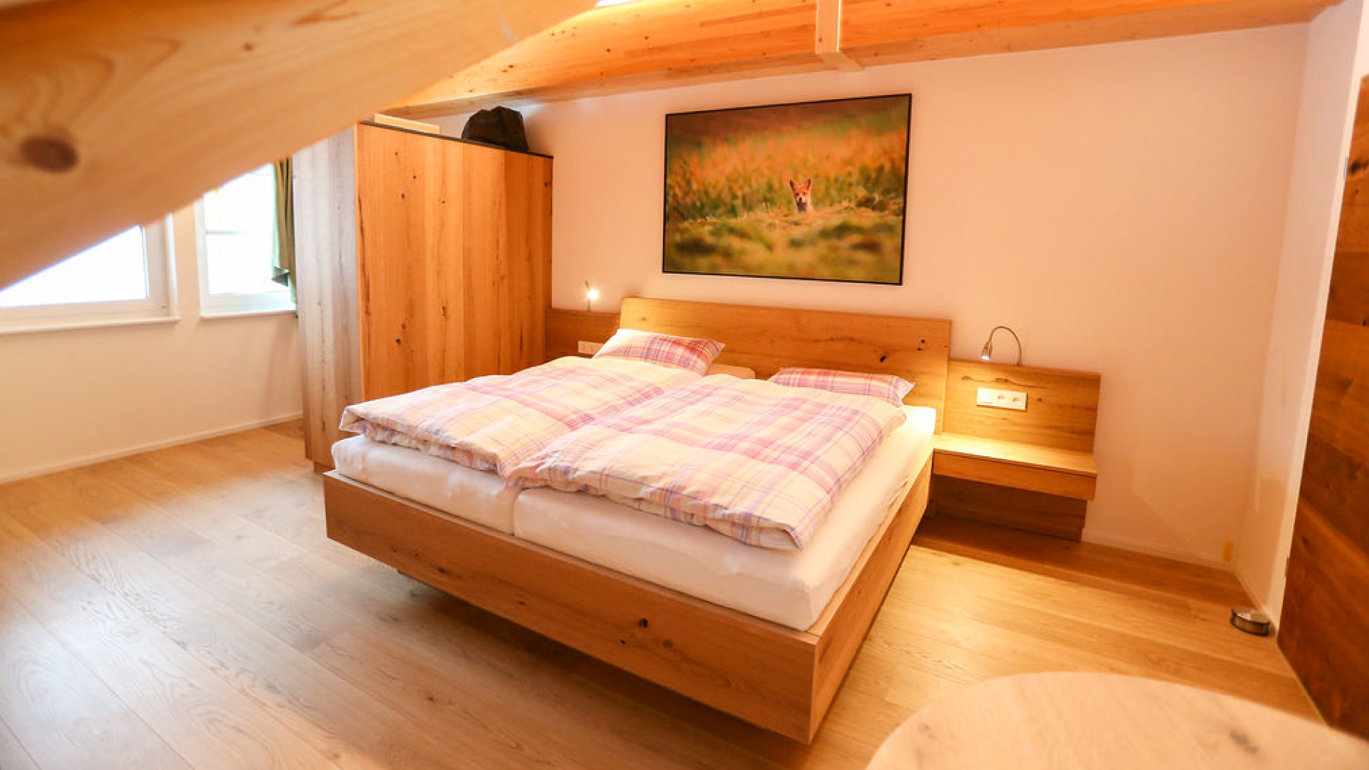 Schlafzimmer mit Holzbetten, Wandbild eines Fuchses in einem Feld, Holzregal, Nachttisch mit Leselampe, helle Wände, Fenster mit grünem Vorhang, Holzboden.