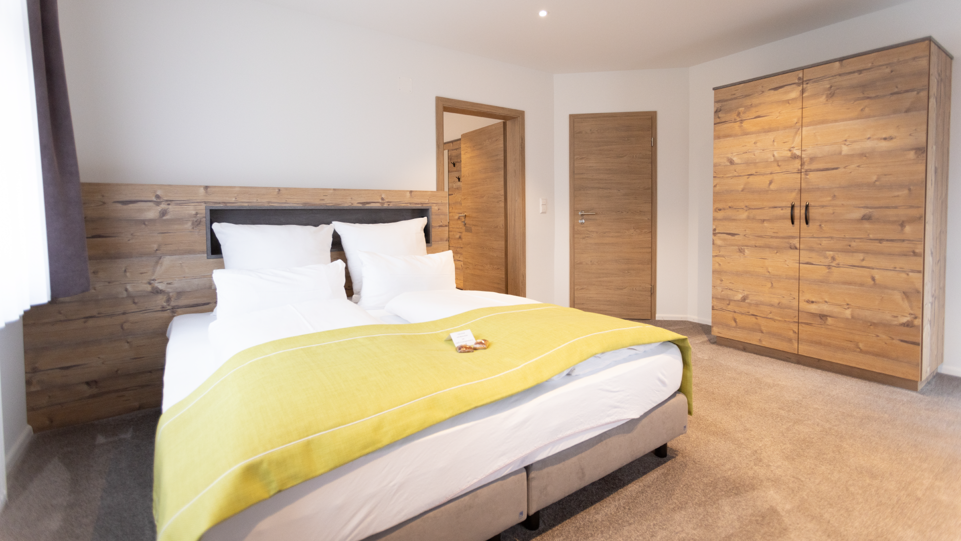 Schlafzimmer mit Holzdetails, weißem Bett mit grüner Decke, großen weißen Kissen, Holz-Schrank, Holztüren, Teppichboden, weiße Wände.