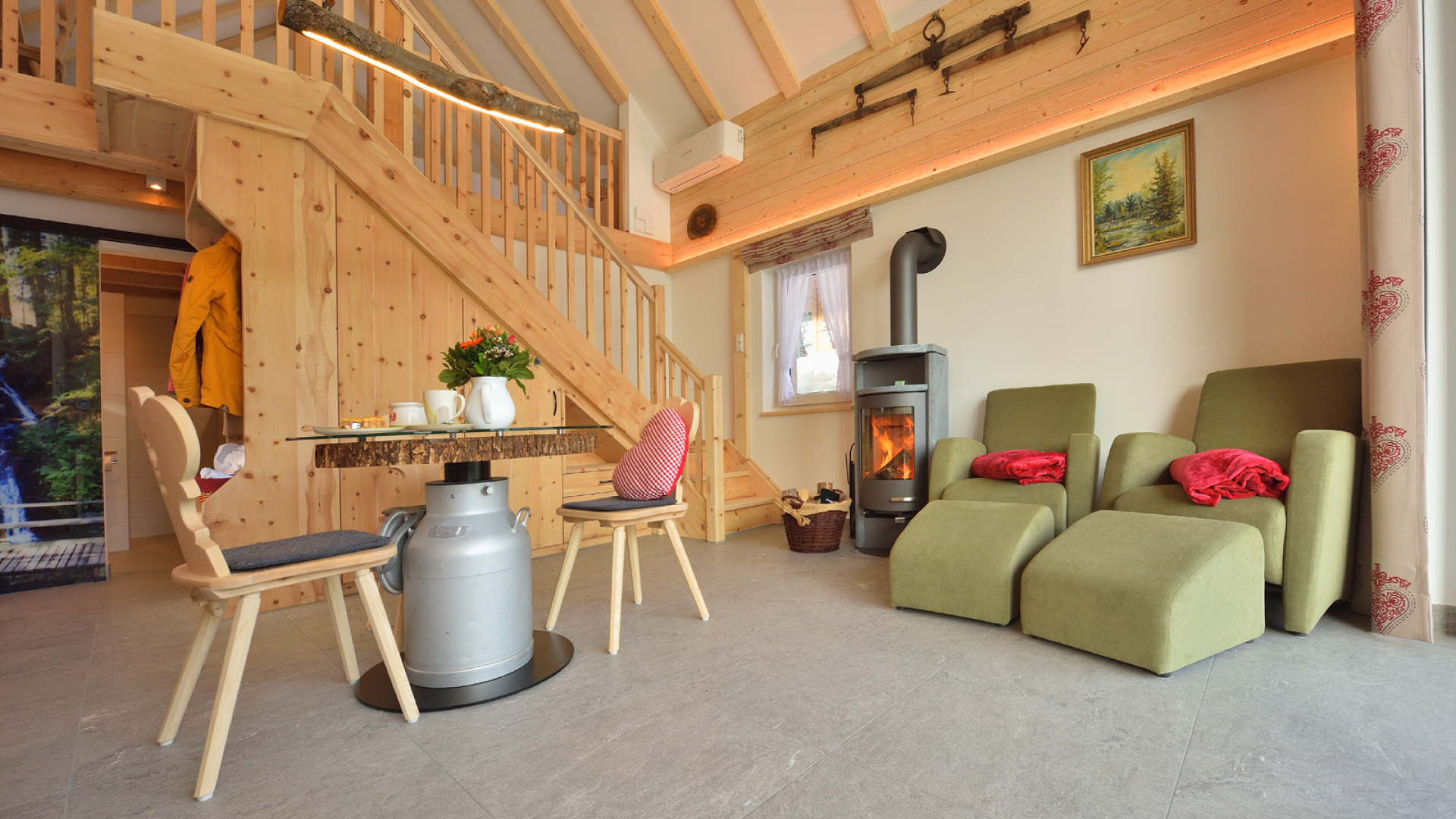 Wohnzimmer mit Kamin, grünen Sesseln, Treppe aus Holz, kleiner Tisch, Stühlen und Dekorationsartikeln.