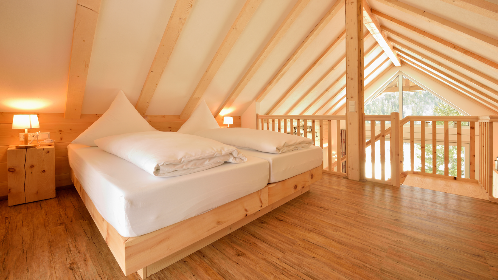 Schlafzimmer in einer Dachgeschosswohnung mit Holzmöbeln, zwei Betten, Nachttischen mit Lampen, Holzfußboden, Dachbalken und Fenstern mit Blick auf Bäume.