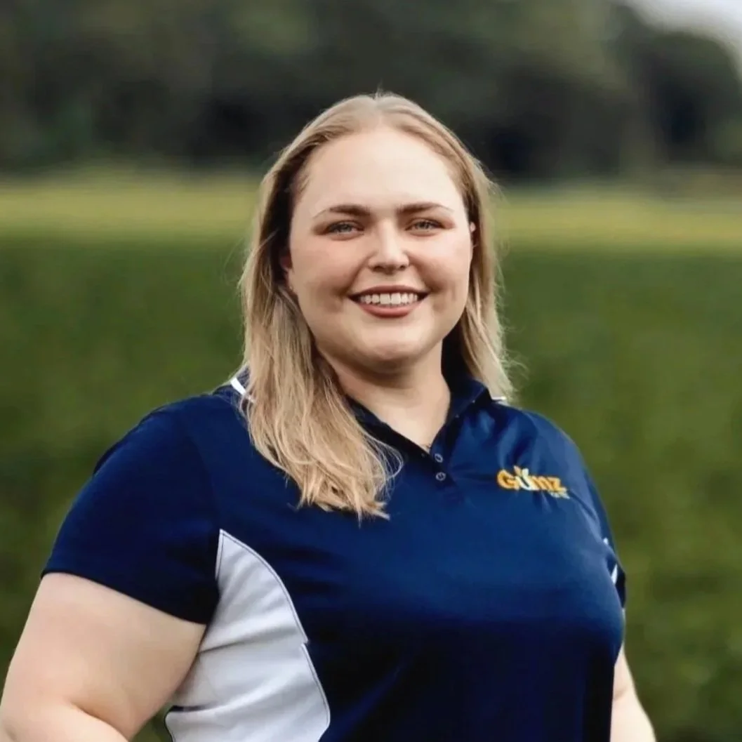 A smiling woman with blonde hair standing outdoors near a lake or pond, wearing a navy blue sports shirt with white accents.