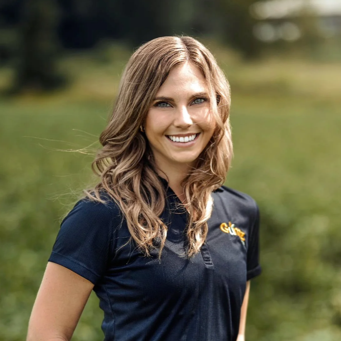 Smiling woman with wavy brown hair wearing a navy blue polo shirt, outdoors with blurred green background.