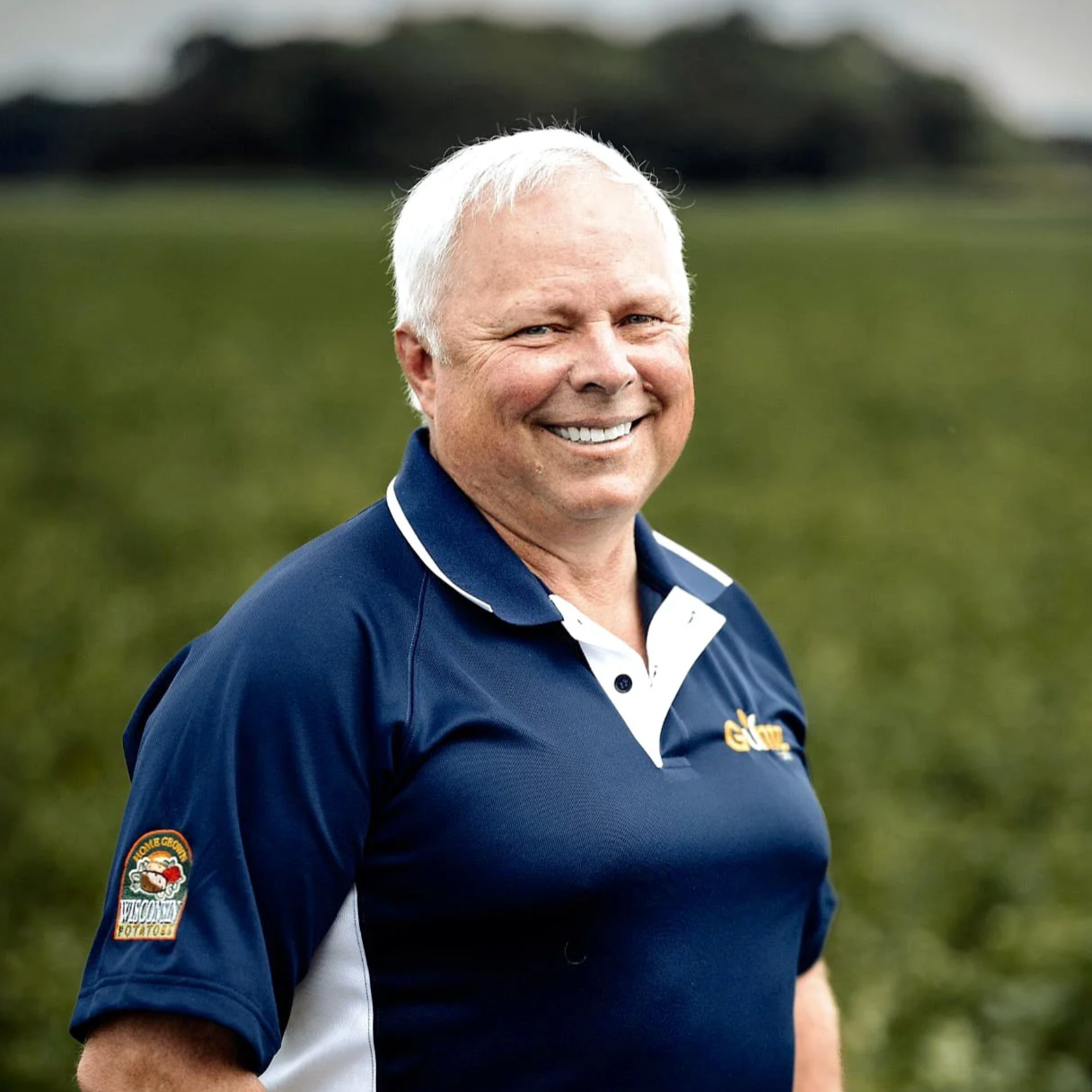 A smiling older man with white hair wearing a navy blue polo shirt with white accents, standing outdoors on a grassy field.