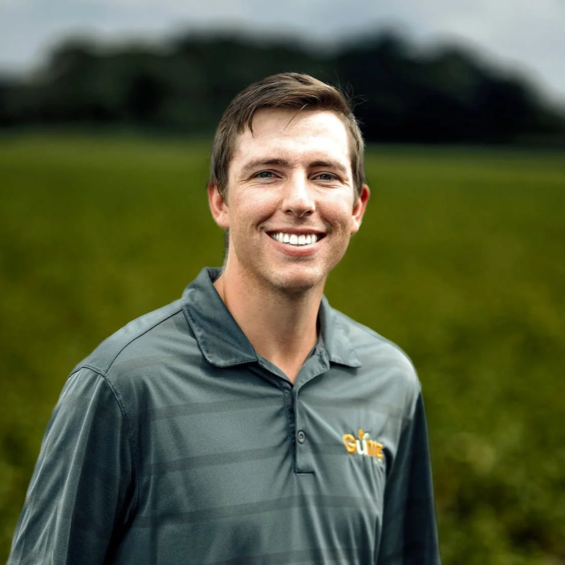 A smiling man in a gray polo shirt stands outdoors in a green field with a blurred background.