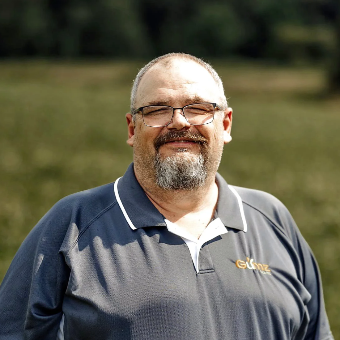A middle-aged man with glasses and a beard, smiling outdoors near a body of water, wearing a dark sports shirt with white trim and a logo on the chest.