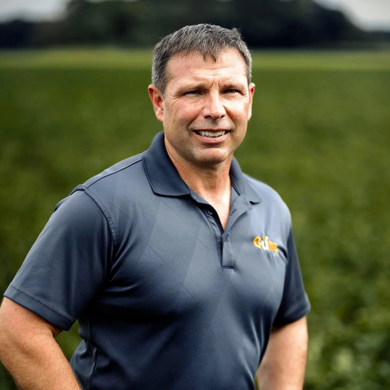 A middle-aged man with short brown hair, wearing a dark blue polo shirt with a logo, standing outdoors with greenery in the background.