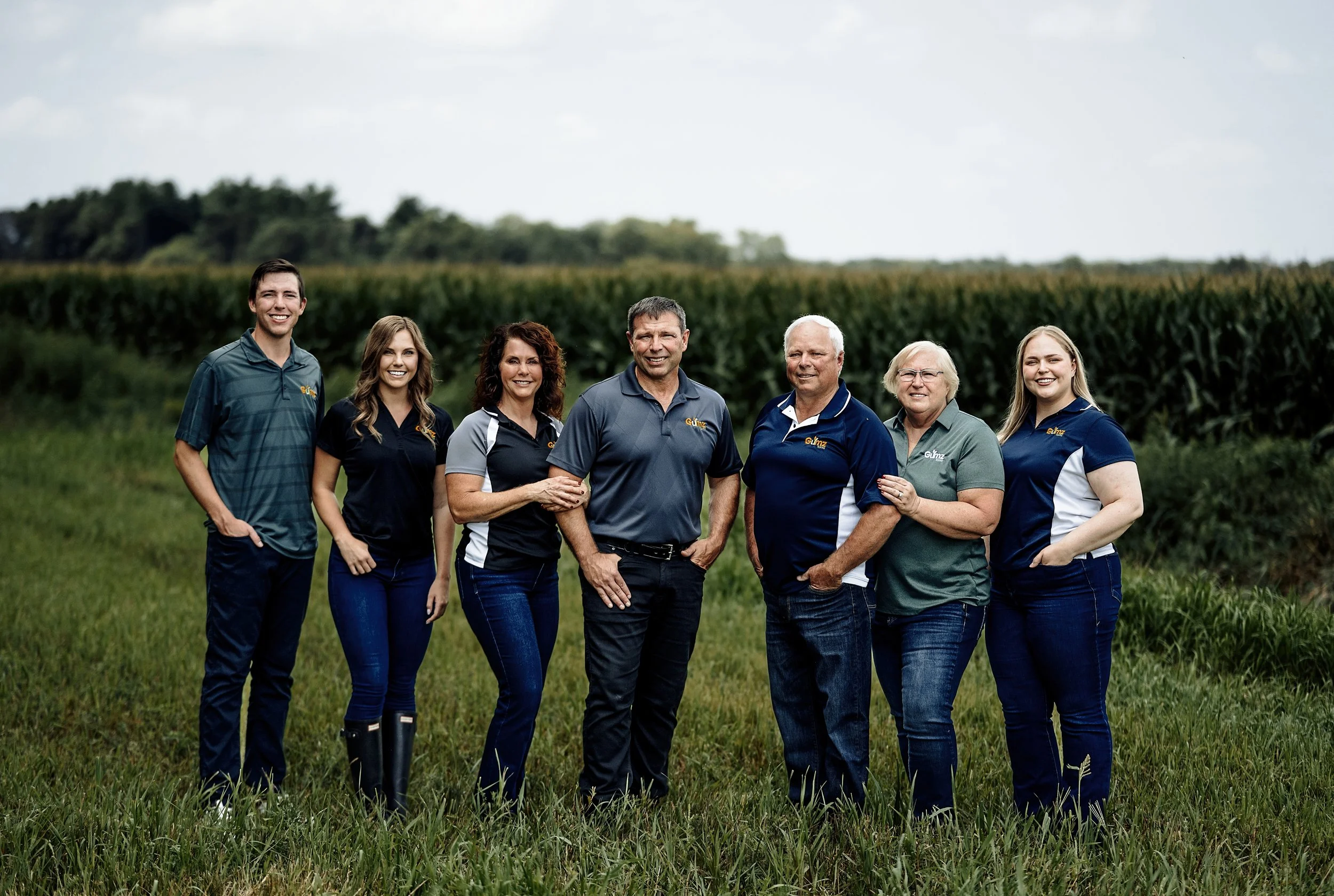Group of seven people standing in a grassy field with a large cropfield in the background, smiling at the camera.