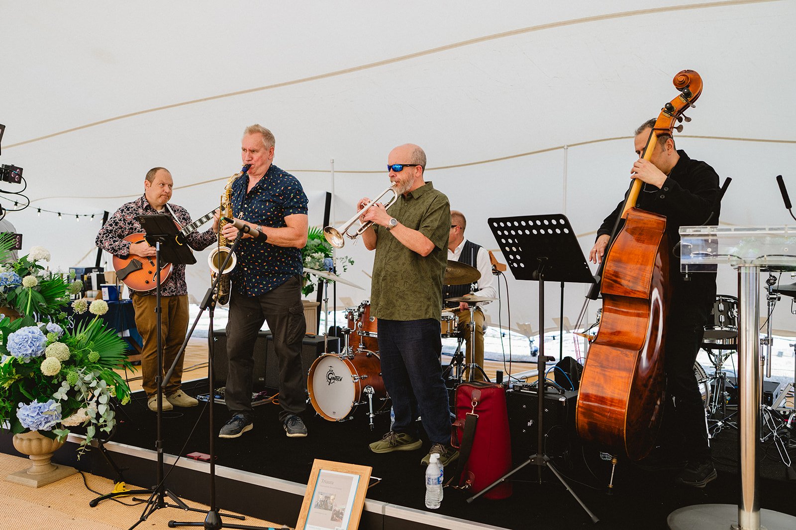 A band of five musicians performing under a large tent, with flowers and decorative items around the stage. They are playing guitar, saxophone, trumpet, drums, and double bass.