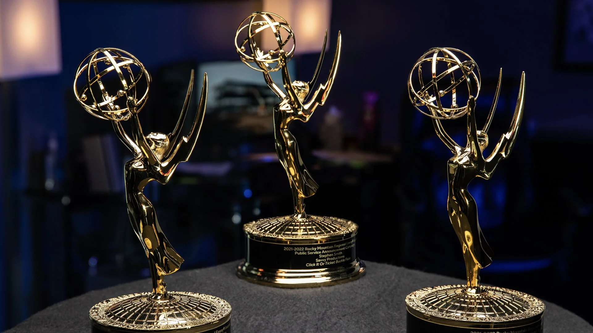 Three gold Emmy-style awards with a stylized human figure holding a globe, displayed on a dark surface with a blurred background.