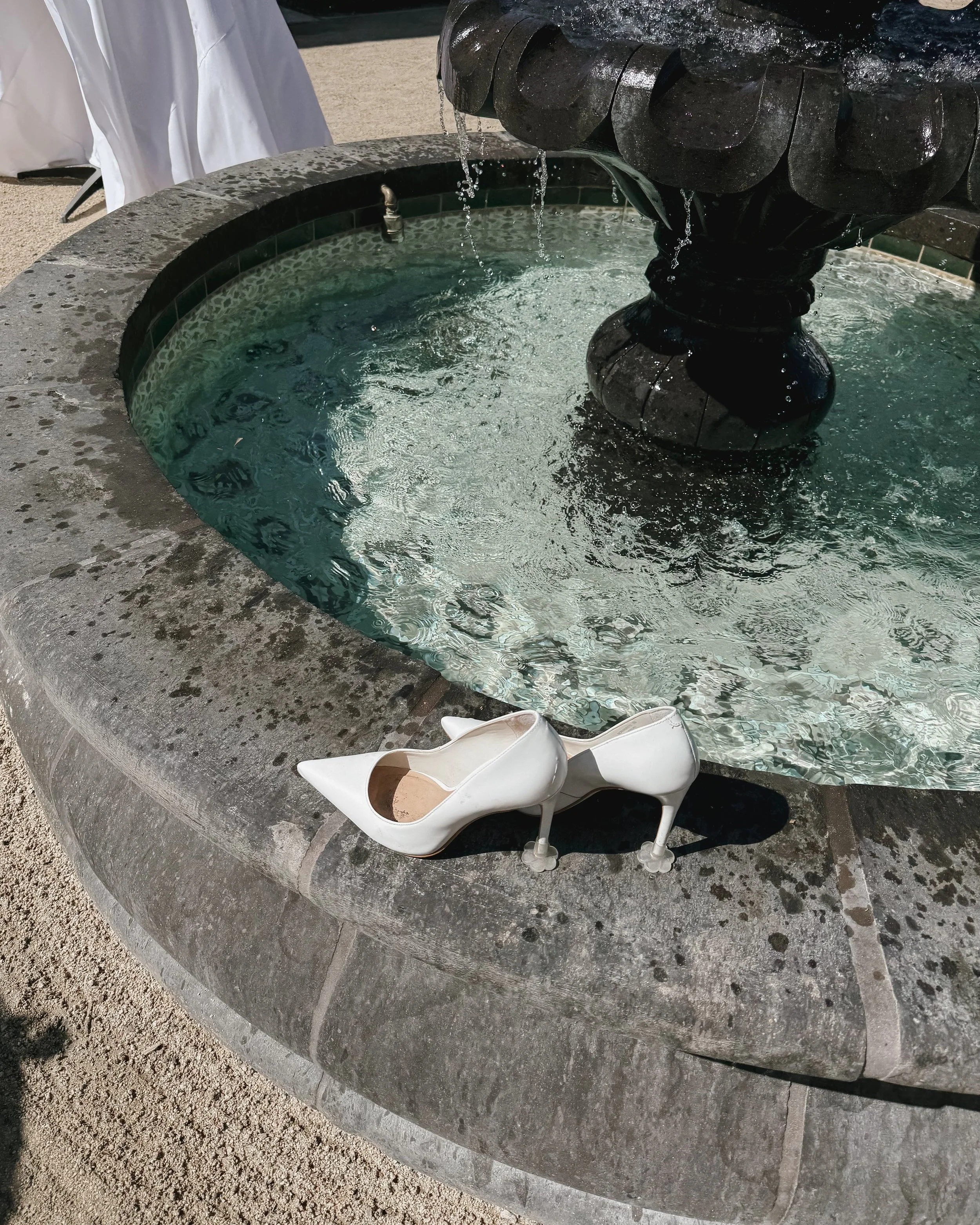 A pair of white high-heeled shoes placed on the edge of a stone fountain with water, in an outdoor setting.