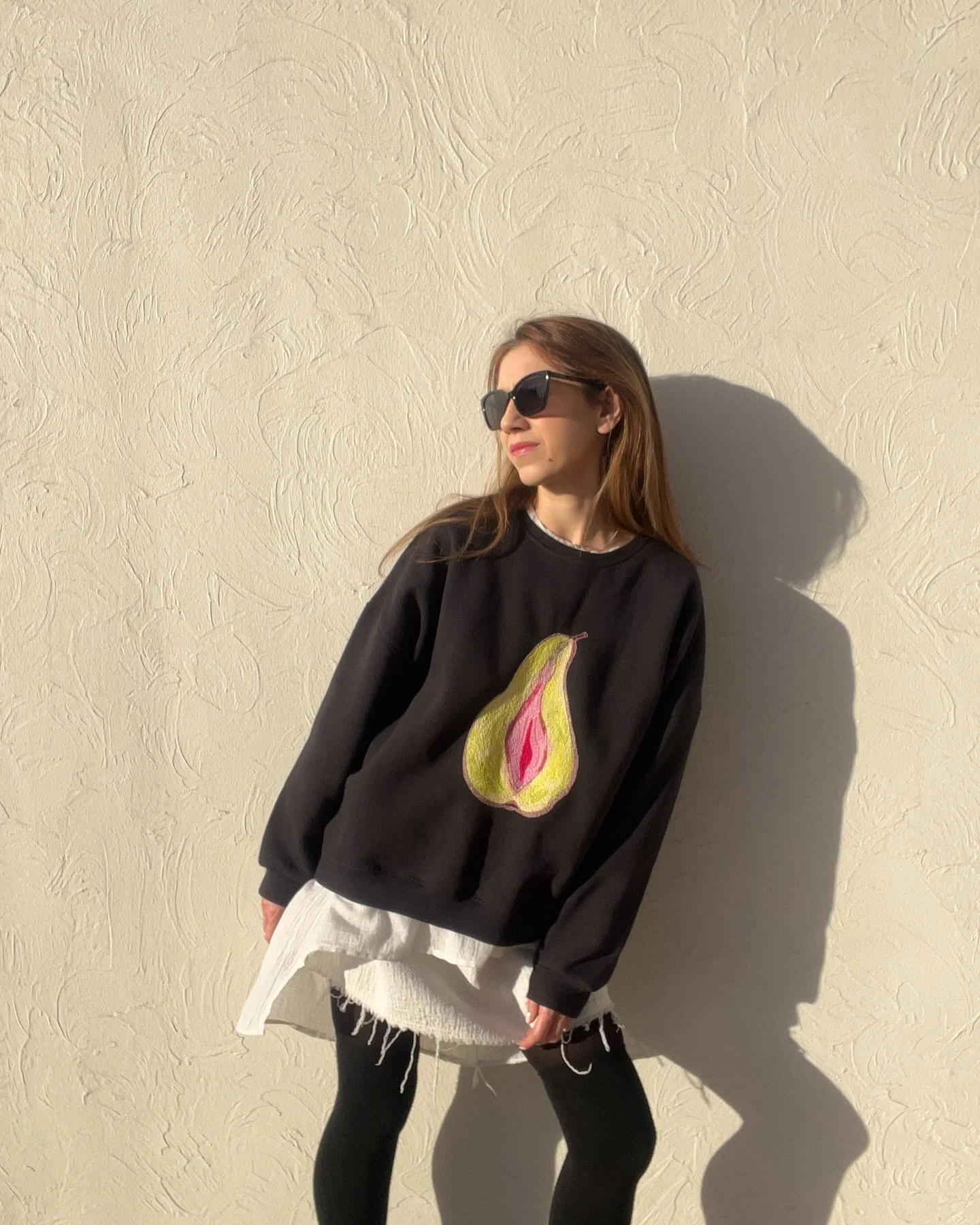 Woman wearing black sweatshirt with a pear design, white shirt underneath, black leggings, and sunglasses standing against a textured beige wall with a shadow cast behind her.