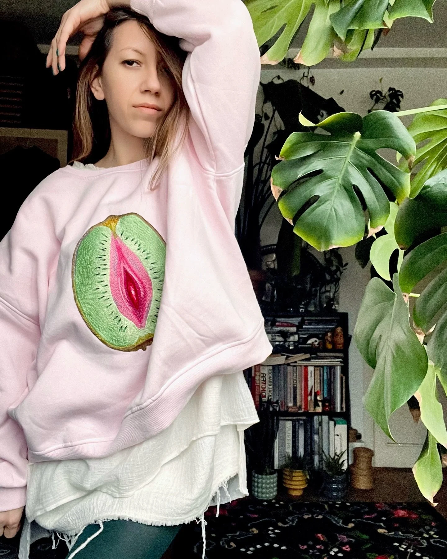 A woman with light brown hair, wearing a pink sweatshirt with an embroidered kiwi fruit featuring a pink interior, poses with her arm raised above her head. She is in a room with large green houseplants, black bookshelves filled with books, and other decorative items in the background.