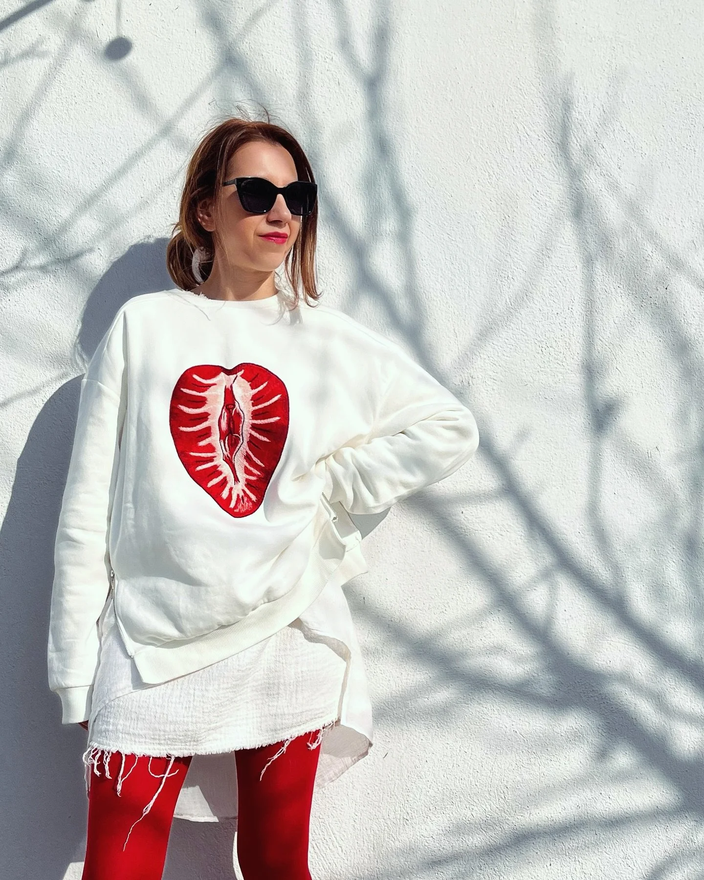 Woman with dark sunglasses, wearing a white sweater with a red lips design, red leggings, standing against a textured white wall with shadows.