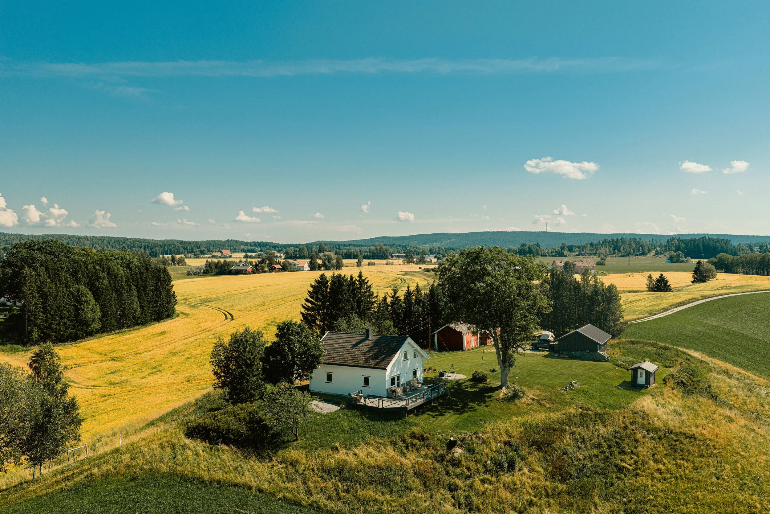 Landlig utsikt med hvitt hus, grønne trær, gressletter og gule åkre under en klar blå himmel.