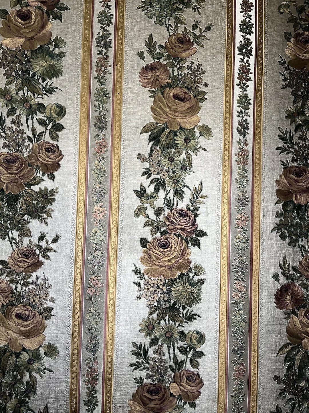 Close-up of a vintage floral wallpaper pattern featuring vertical stripes with large roses, daisies, and smaller flowers in muted tones of beige, green, and pink.