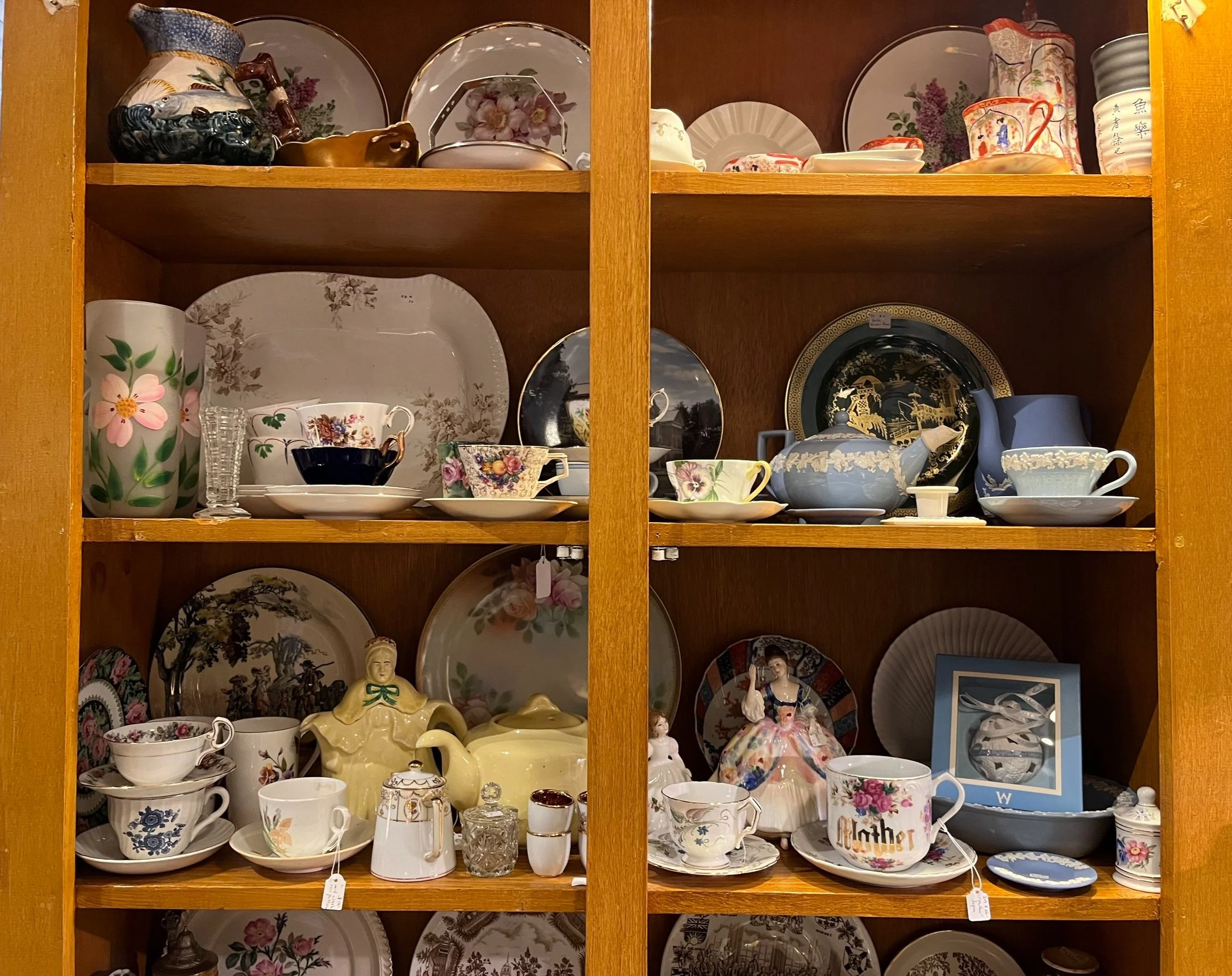 A wooden display cabinet filled with various decorative porcelain and ceramic dishware, including plates, teapots, cups, and figurines, with floral and traditional Asian designs.