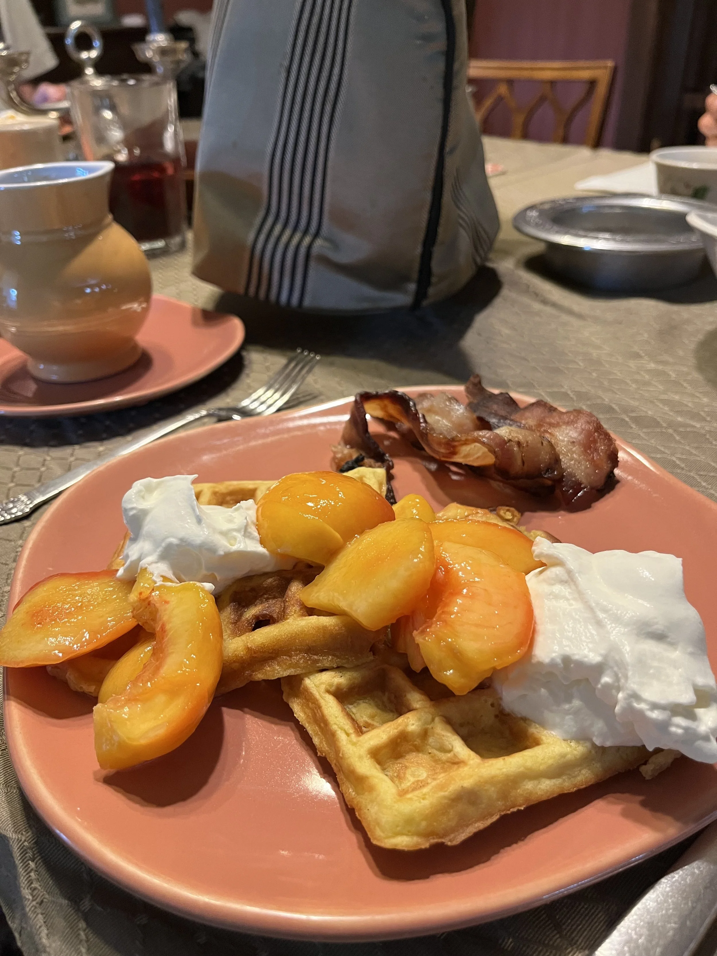 A breakfast plate with waffles topped with peaches and whipped cream, a piece of crispy bacon, and a waffle with some syrup. The table has a cup and saucer, various dishes, and utensils.