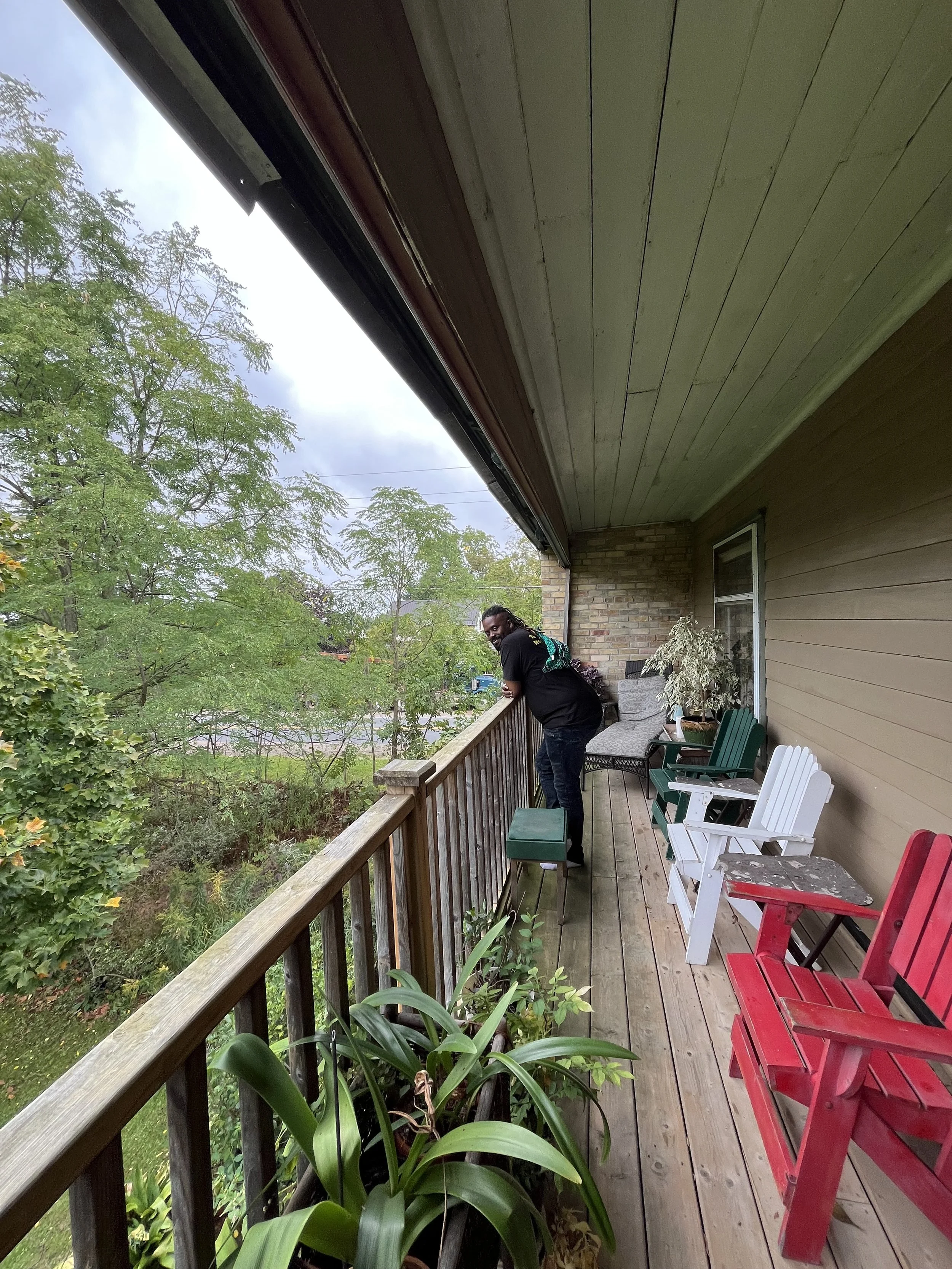 A man leaning on a wooden balcony railing, looking at the camera, with green chairs, a white chair, a red chair, and potted plants on the porch, overlooking a leafy yard with trees.
