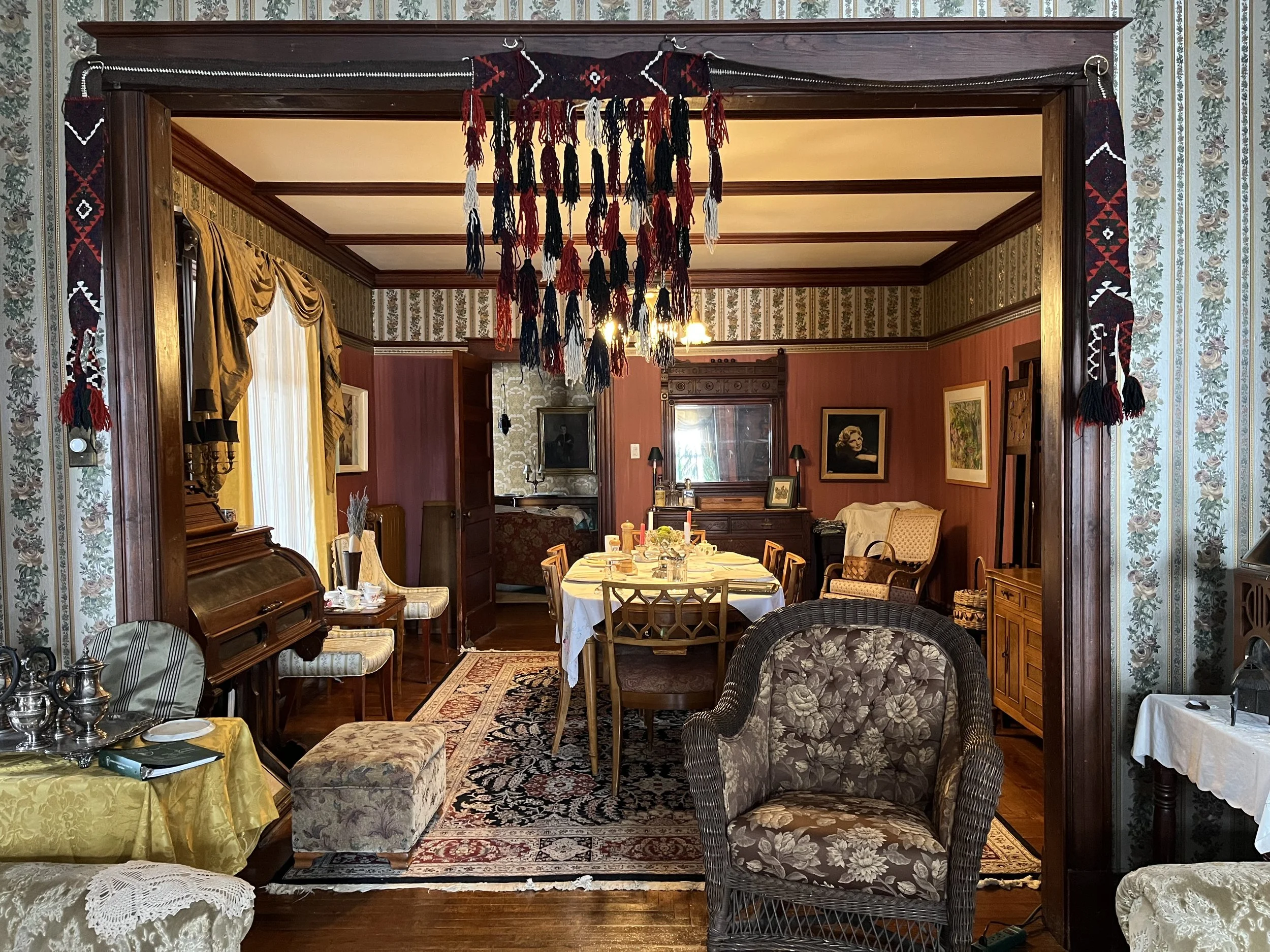 A vintage-style dining room with a round table set with candles and dishes, surrounded by chairs and upholstered furniture. The room has wooden trim, floral wallpaper, and a large tapestry hanging from the ceiling.