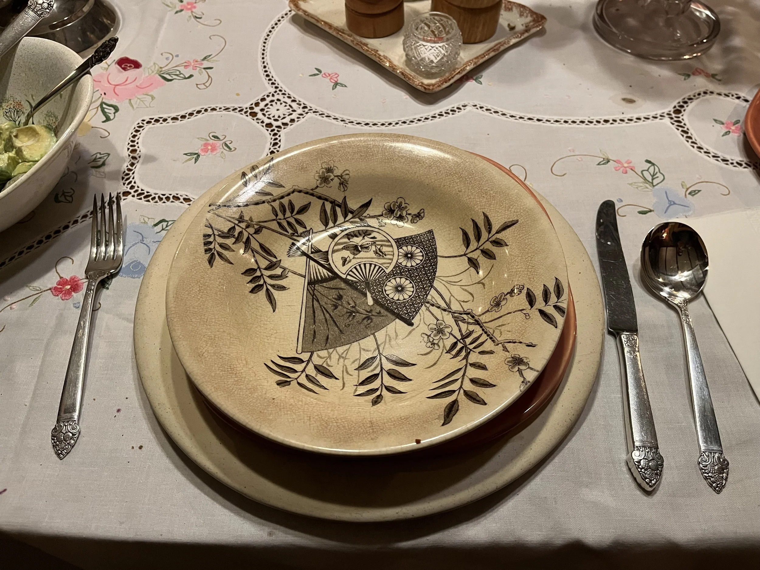 A decorative plate with a black floral design stacked on a beige plate, placed on a table with silverware on either side, and a tablecloth with painted floral patterns.