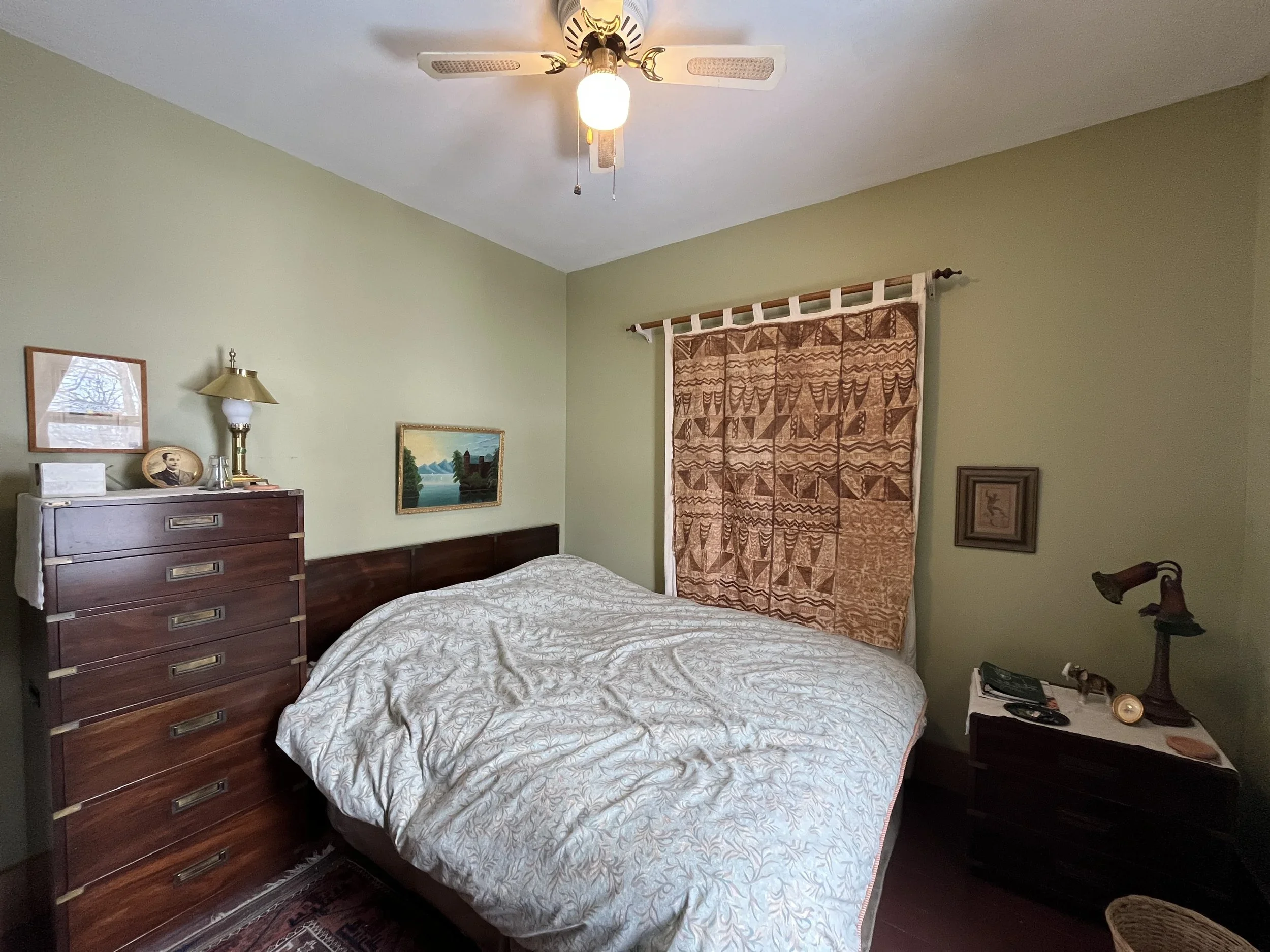 A bedroom with a bed covered with a striped quilt, two pillows, a wooden dresser with a lamp, picture, and glasses on top, a green wall with a framed landscape painting, and a window with patterned curtains.