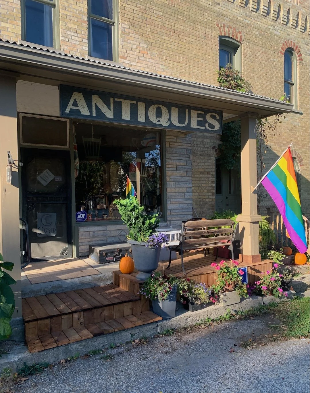 Front of an antique shop with a sign 'ANTIQUES', decorated with rainbow flags, pumpkins, potted plants, and flowers outside.