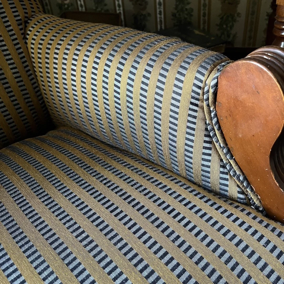 Close-up of a vintage-style sofa with striped fabric upholstery in gold, black, and blue tones, featuring a rolled armrest and wooden accents.