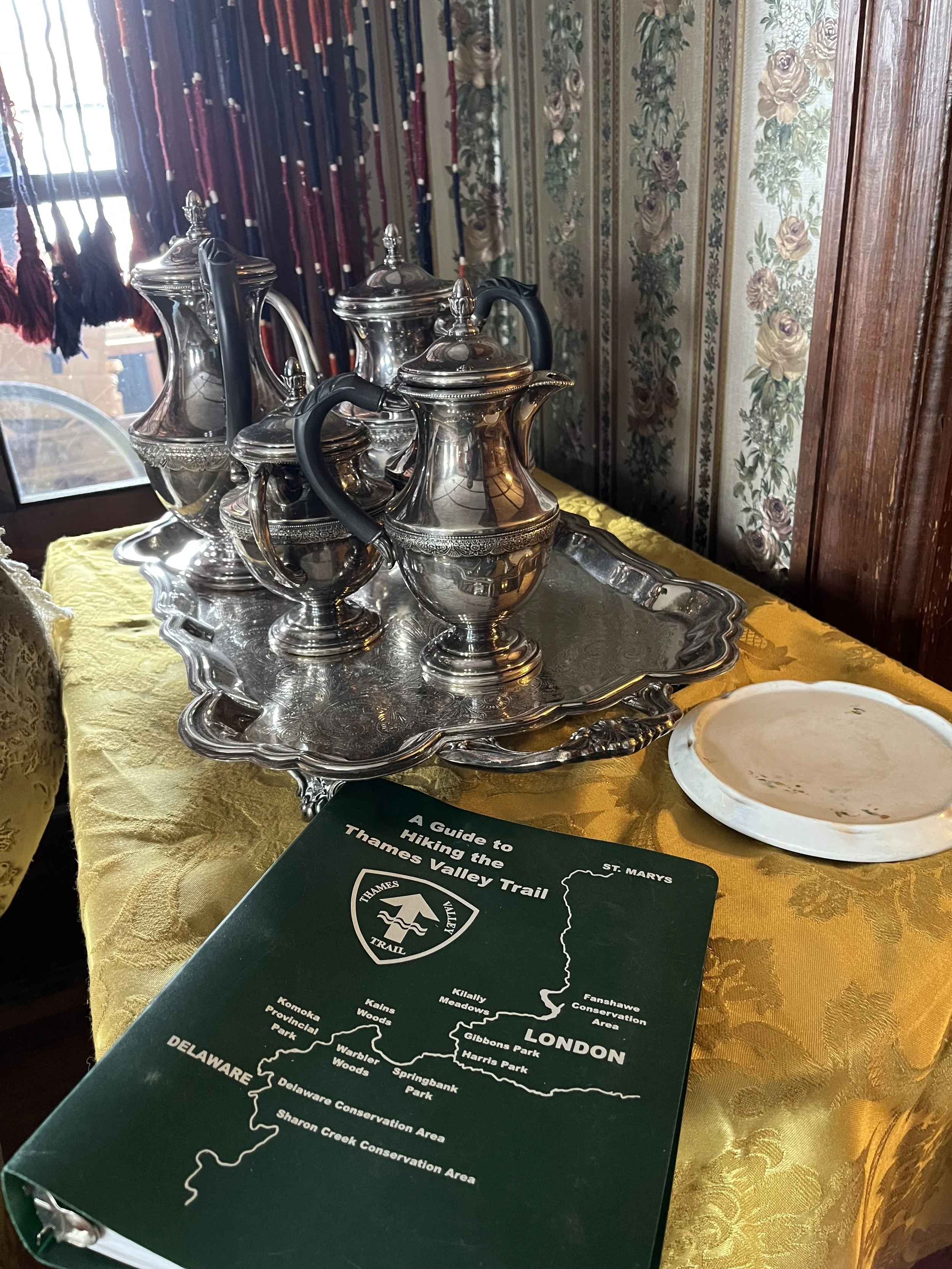 A silver tray with three silver teapots and a cream jug on a yellow tablecloth, with a plate, a registration pamphlet titled 'A Guide to Hiking the Thames Valley Trail,' and a window with red and blue curtains in the background.