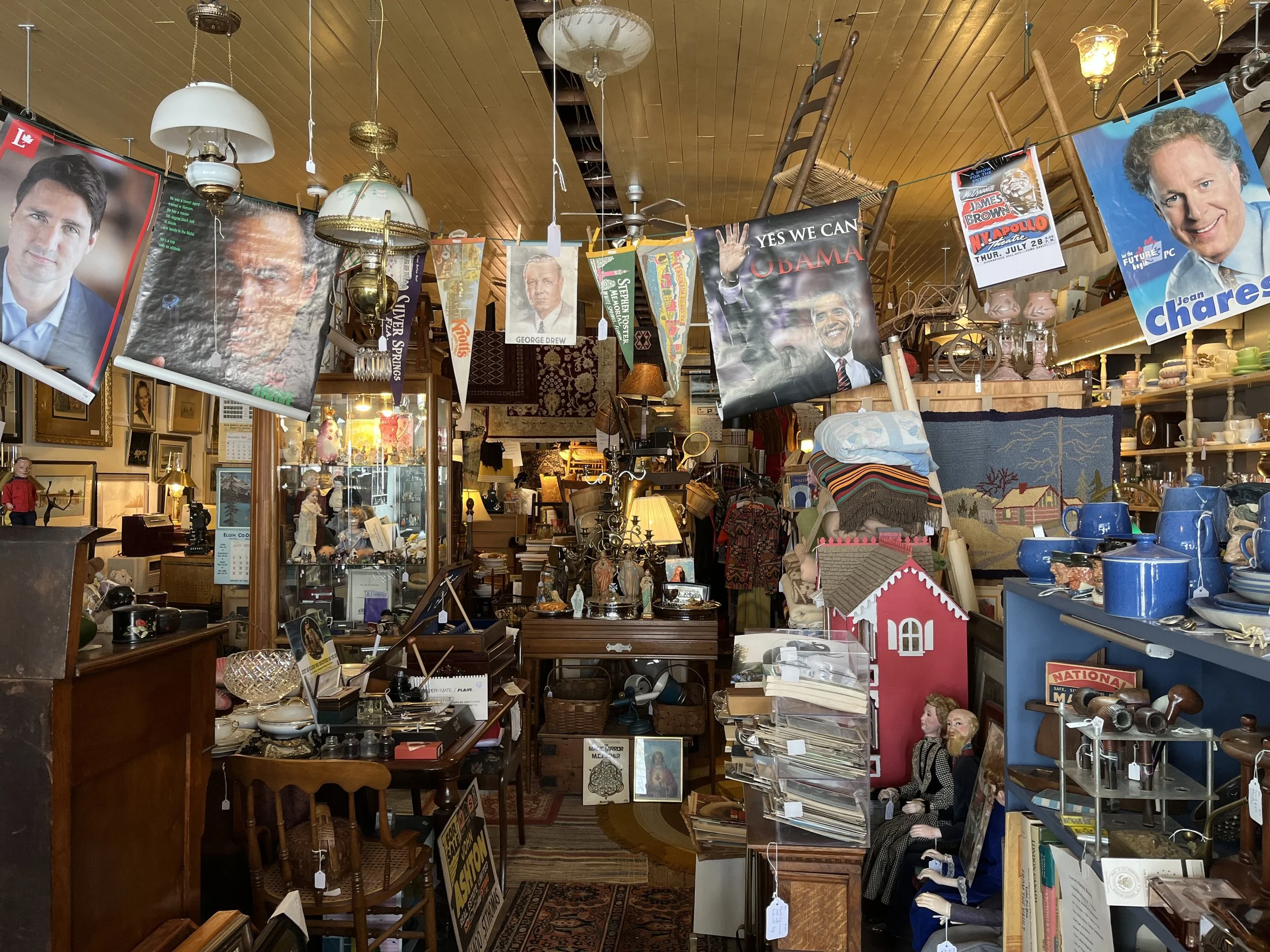 Interior of a vintage thrift shop filled with various items including framed posters, books, dolls, dishes, and decorative objects.