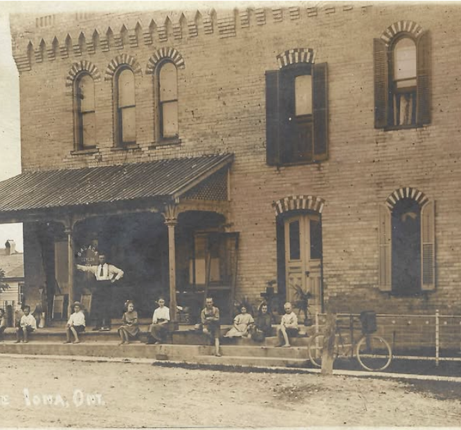 Historical black and white photo of a group of children sitting on a sidewalk in front of a two-story brick building with a porch and open window shutters.