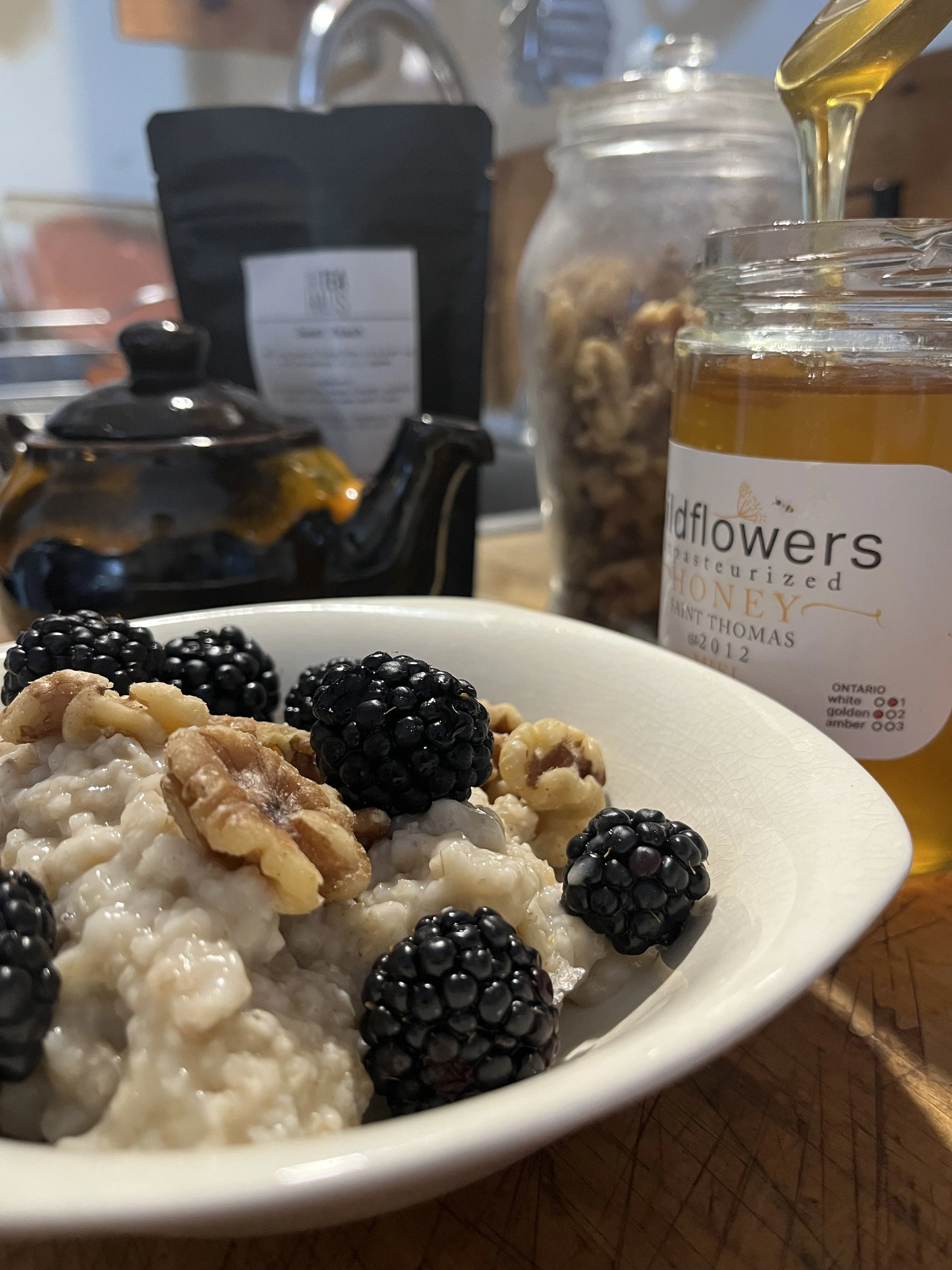 A bowl of cottage cheese topped with blackberries and walnuts, with honey and various jars and a teapot in the background.