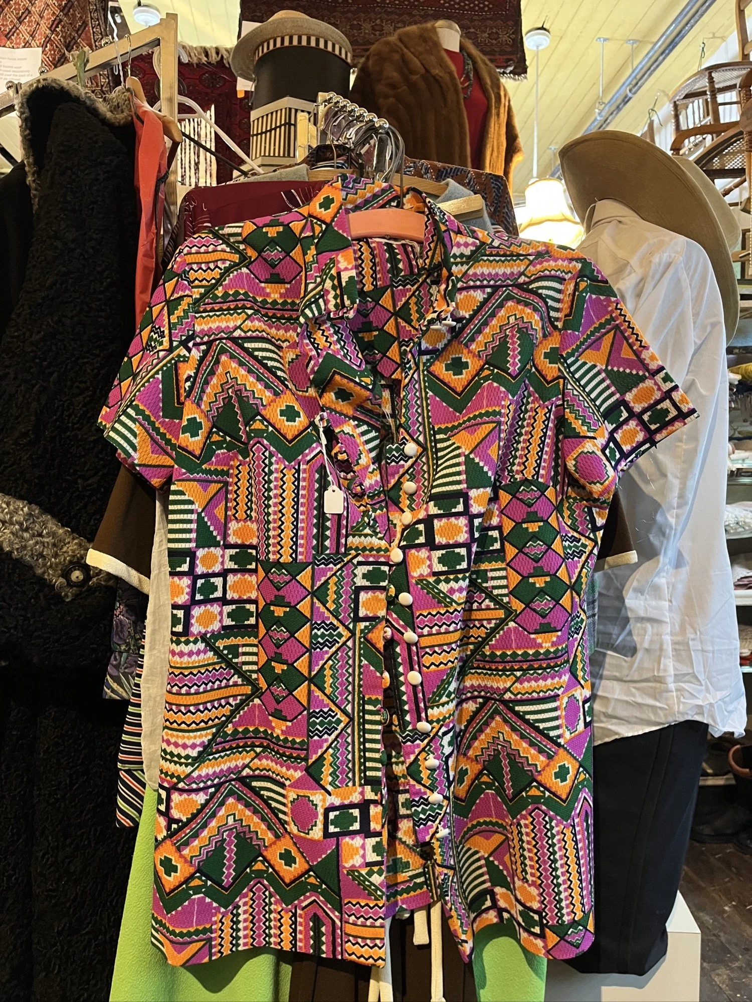 A beige suede tunic with fringe details on the sleeves, displayed on a mannequin with a beaded necklace, in a rustic room with wooden furniture and shelves.