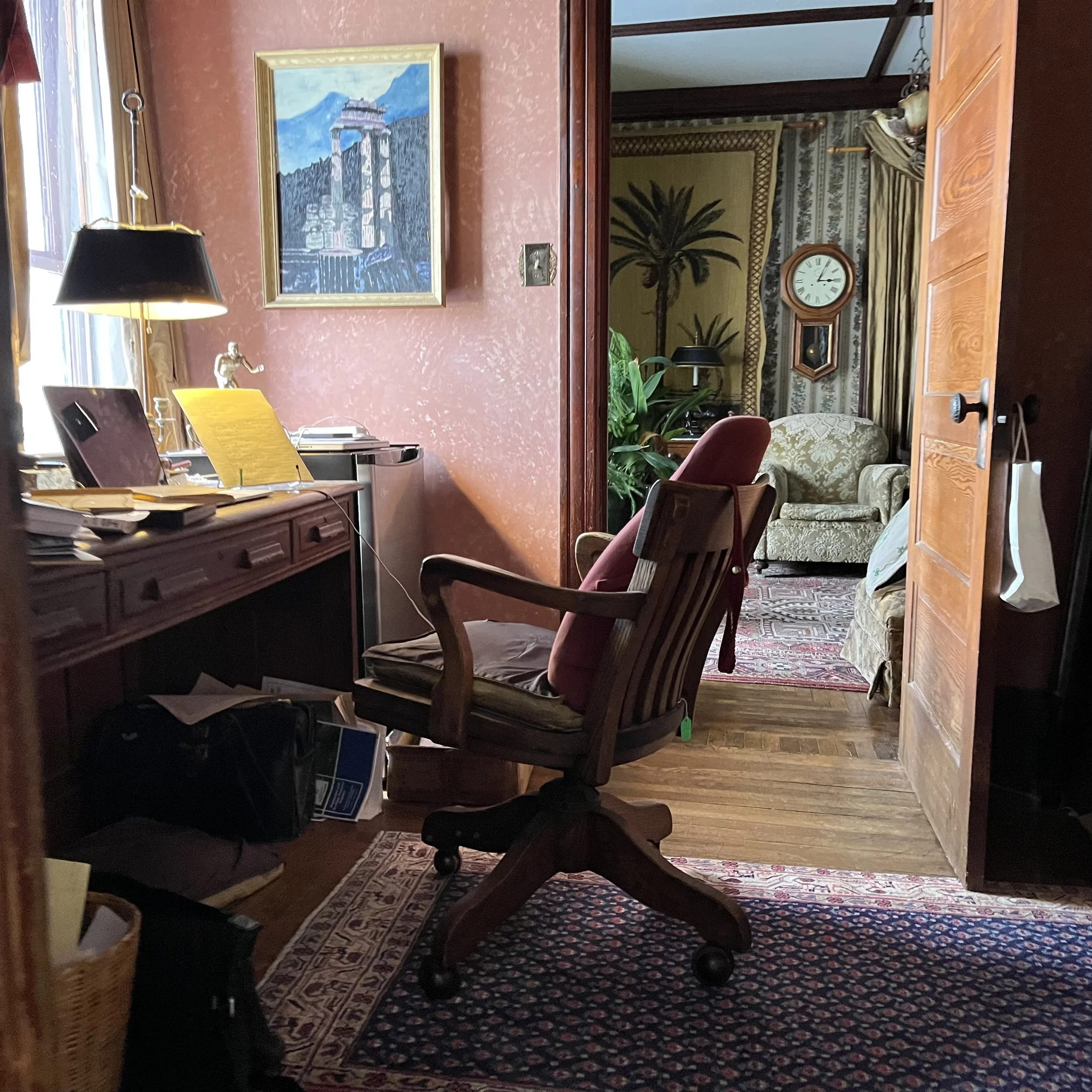 A home office with a wooden desk, a pink armchair, and a cushioned wooden chair. The desk has papers, a lamp, and a desk lamp with a gold base. There is artwork on the pink wall above the desk. Behind the office area, an open doorway leads to a livin