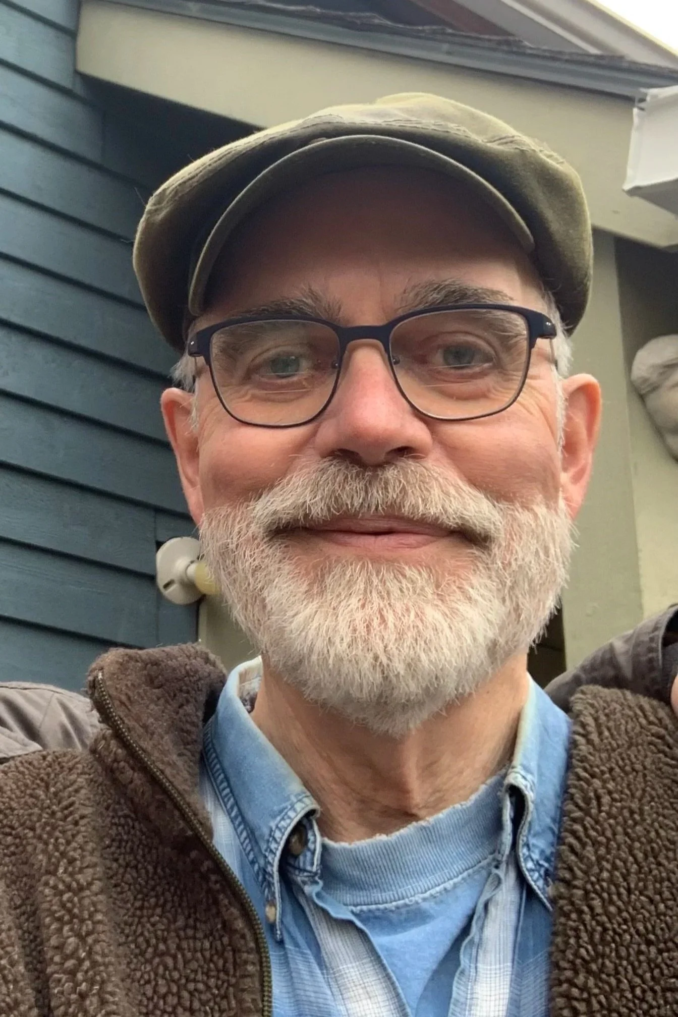The smiling owner Laurence Grant, with glasses, a beard, and a mustache wearing a cap, a plaid shirt, and a brown fleece jacket, standing outdoors near a house with blue siding.
