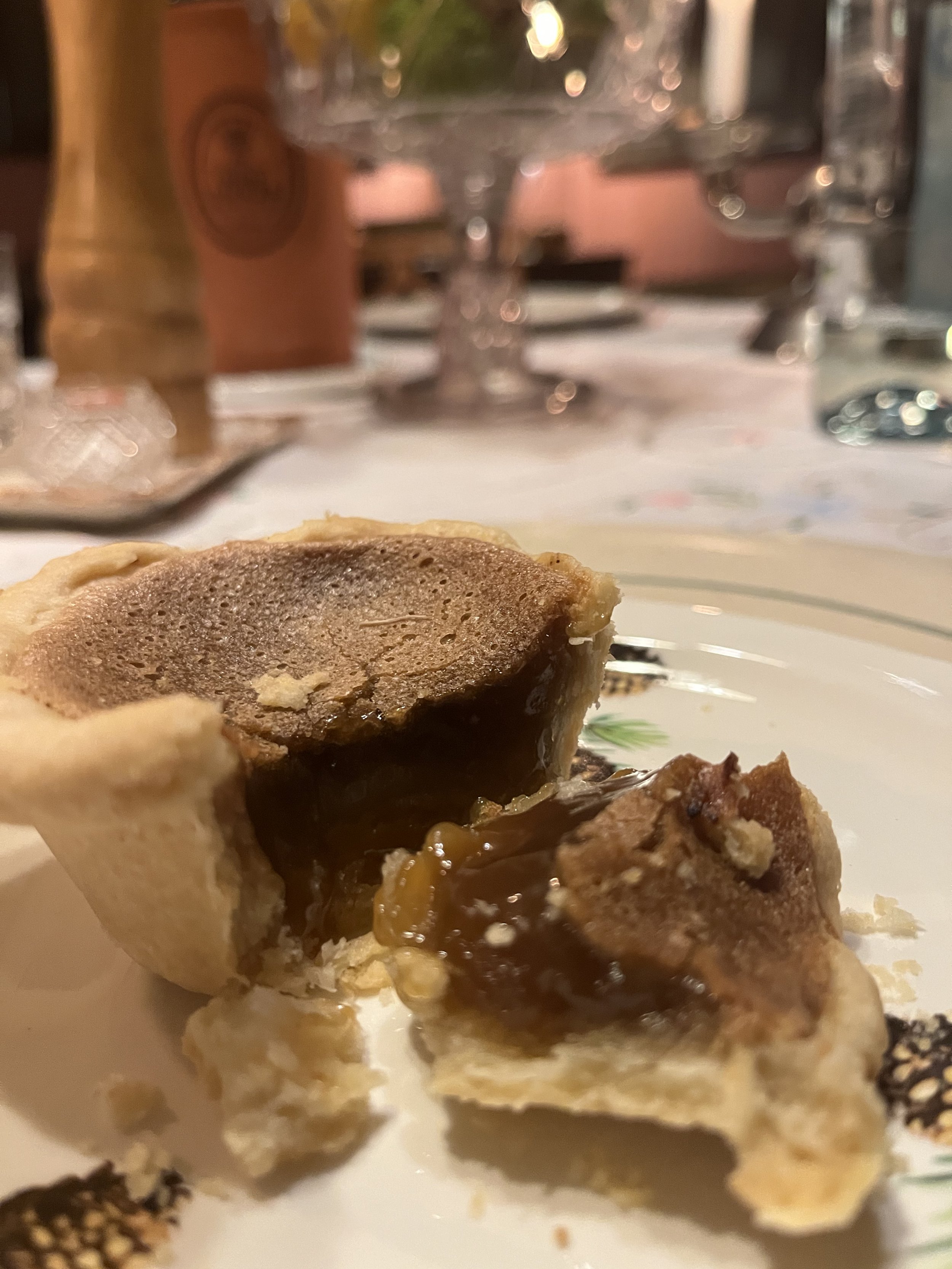Close-up of a partially eaten butter tart with a gooey filling, on a decorative plate, with a blurred dining table in the background.