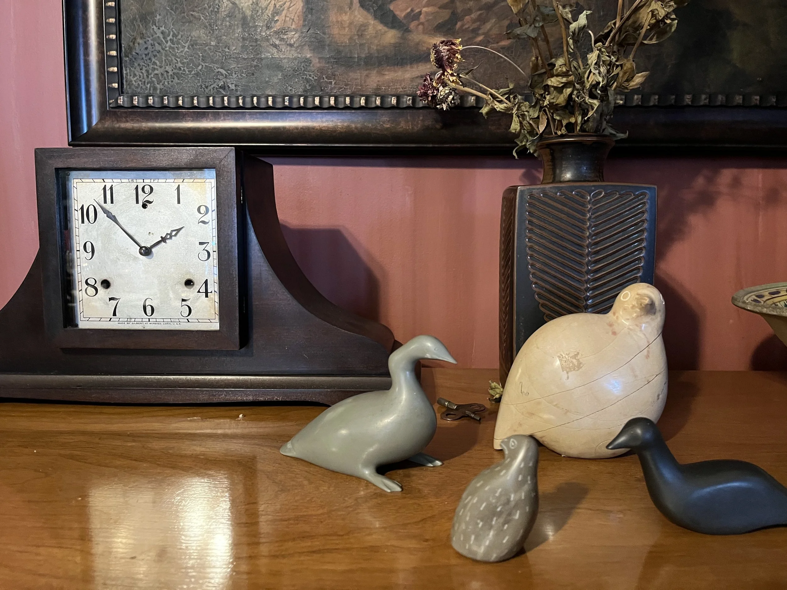 Decorative wooden table with a vintage clock, ceramic bird and animal figurines, dried flowers in a tall vase, and framed artwork on a pink wall.