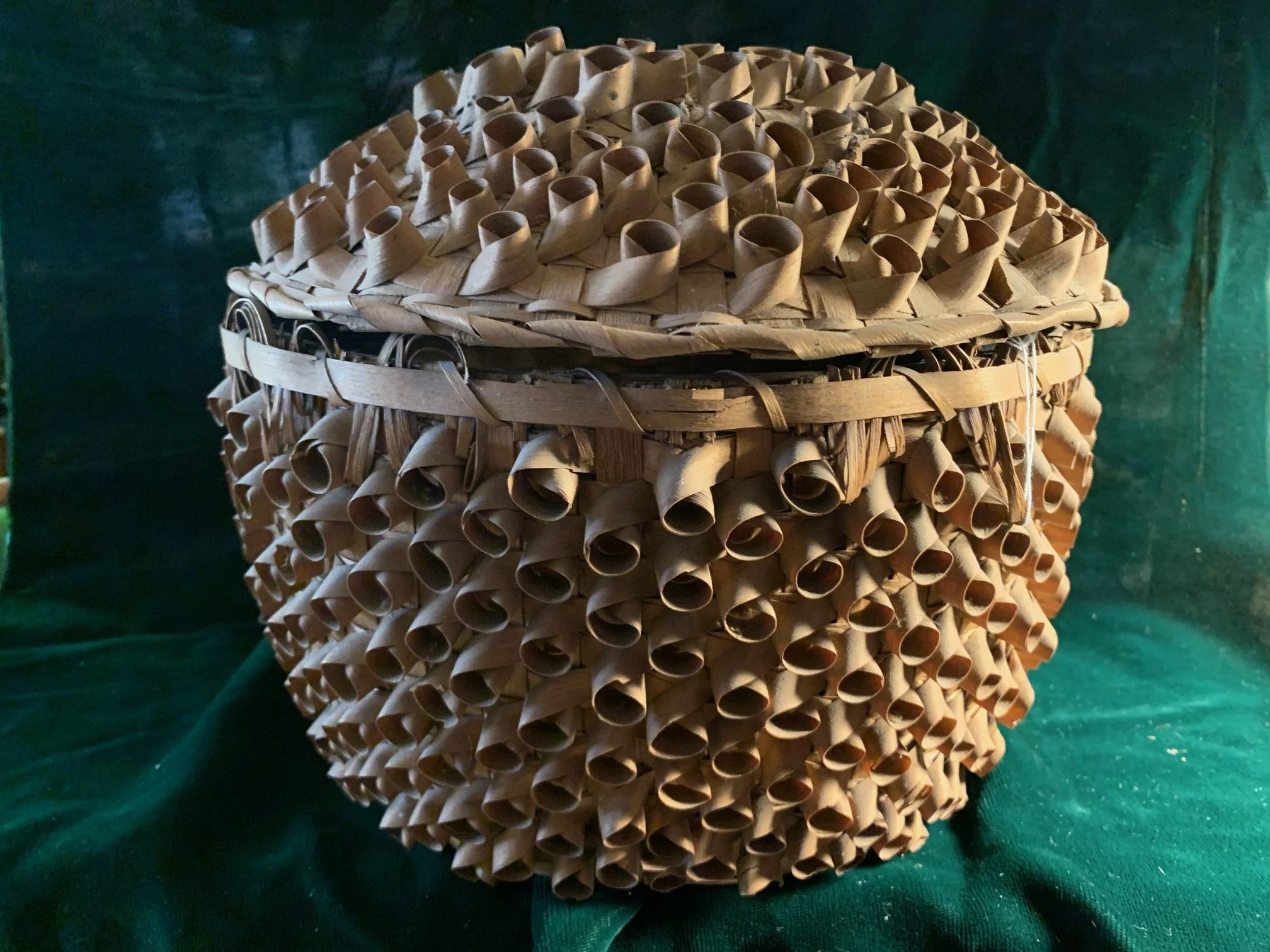 A round, woven basket made of brown natural materials, with a lid. The basket has a textured, spiral pattern and a band across the middle.