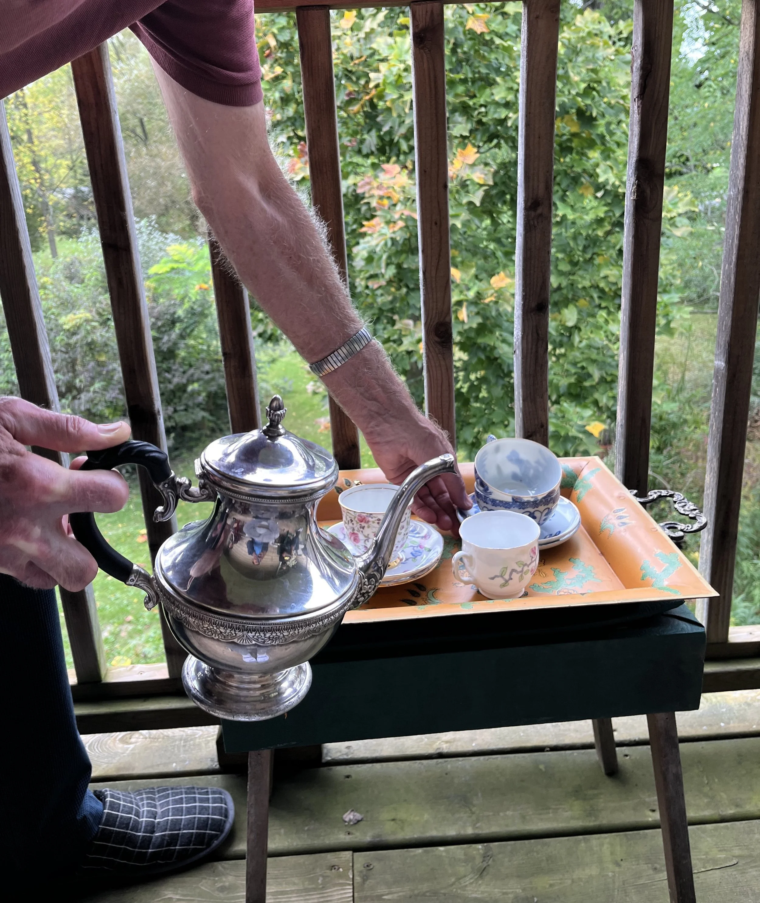 Person pouring tea from a silver teapot onto a tray of assorted teacups and saucers, set on a small black table on a wooden porch with trees in the background.