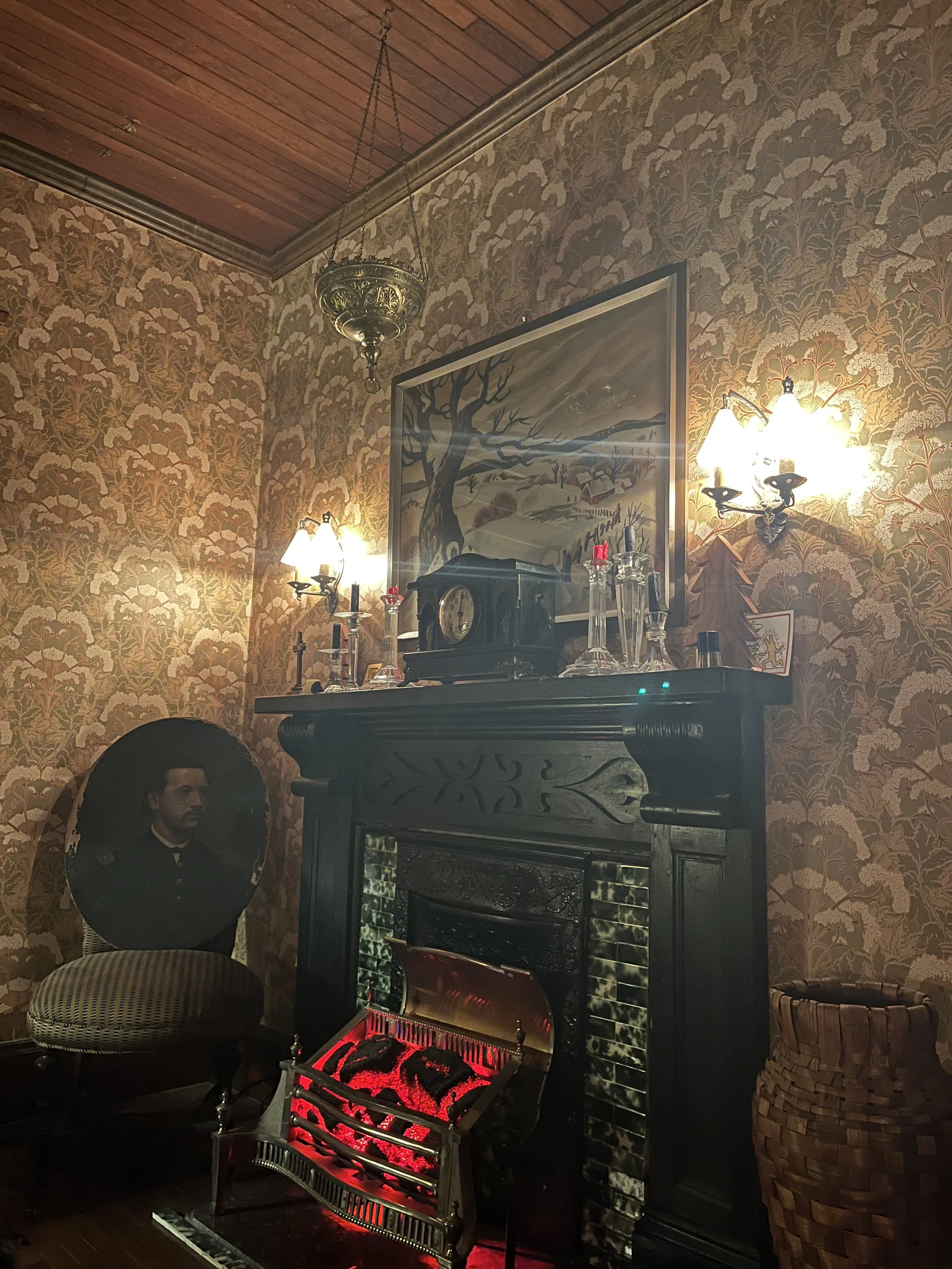 A warm, dimly lit room with vintage decor, including a fireplace with a glowing red fire, a portrait, a painting of a winter landscape, and ornate wall lamps.