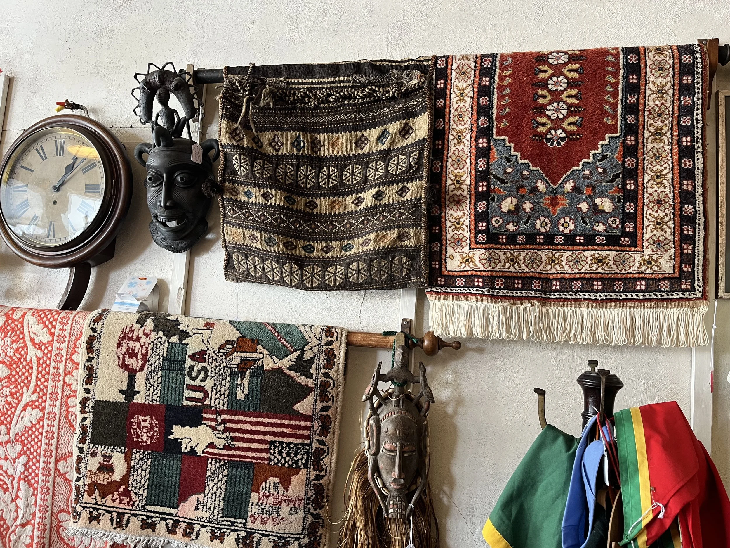 Wall decorated with various woven rugs and masks, including a large round wall clock, a black tribal mask, and other cultural artifacts, with some clothing hanging on hooks.