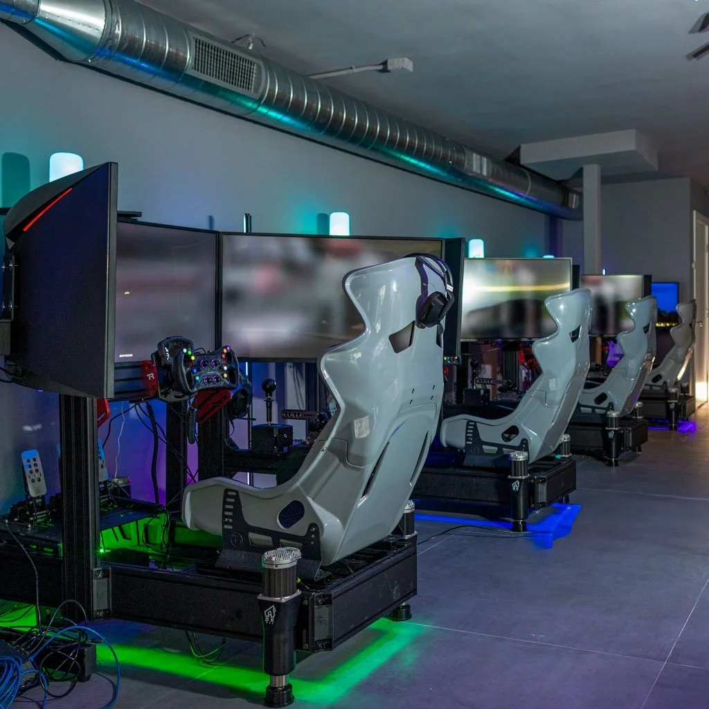 Row of racing simulator setups with large screens, gaming steering wheels, and racing seats in a dimly lit room with colorful LED lighting.