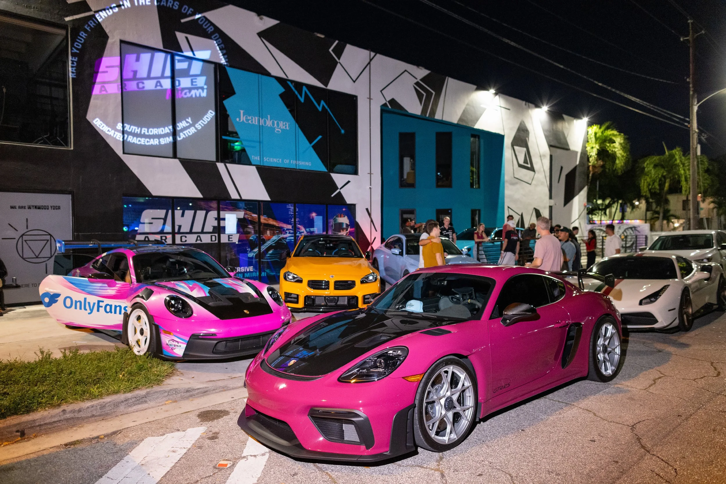 A red and black sports car with a wing spoiler races on a track, featuring Red Bull and Verstappen branding. The background shows blurred greenery and a fence, indicating high speed.