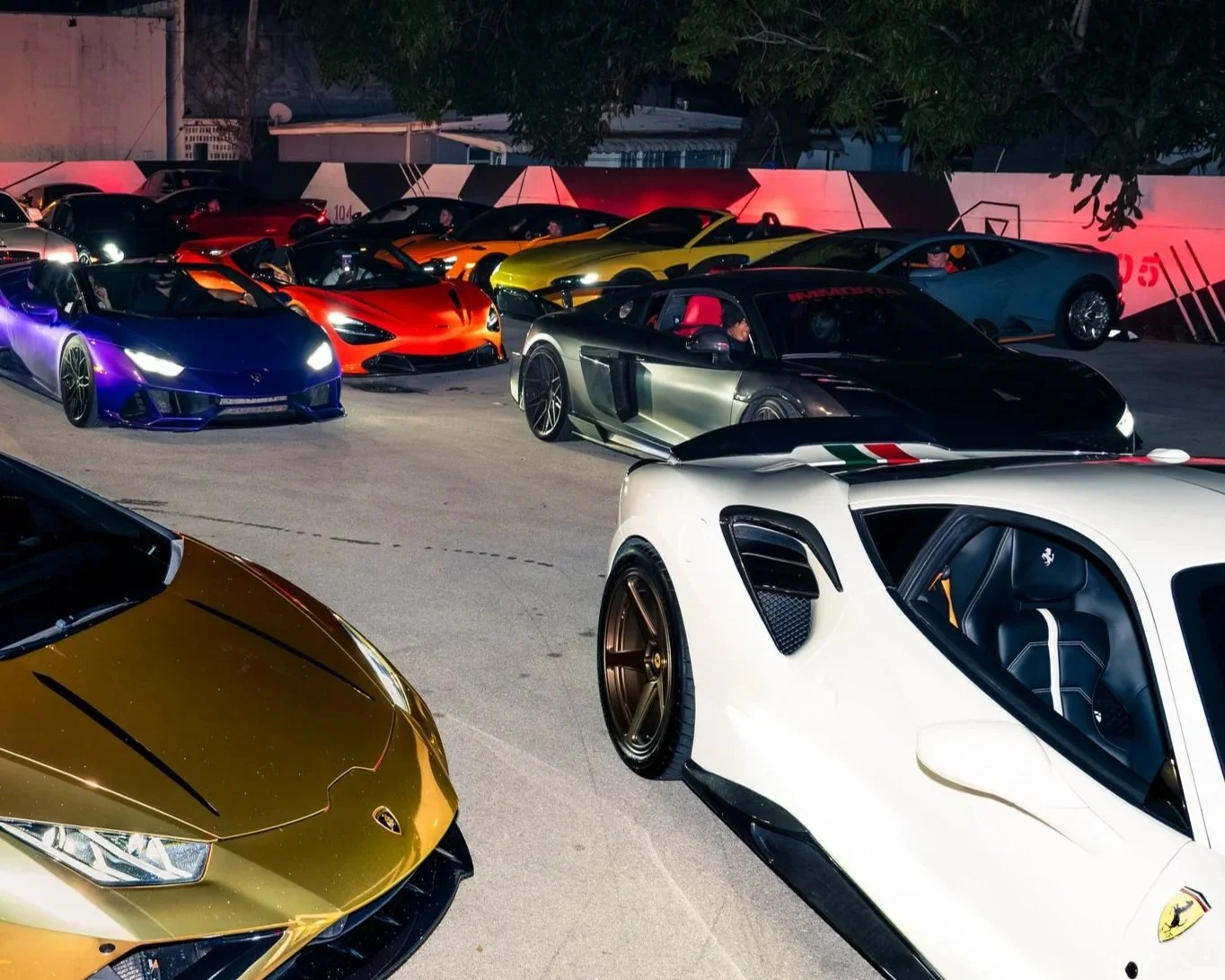 A red and black sports car with a wing spoiler races on a track, featuring Red Bull and Verstappen branding. The background shows blurred greenery and a fence, indicating high speed.