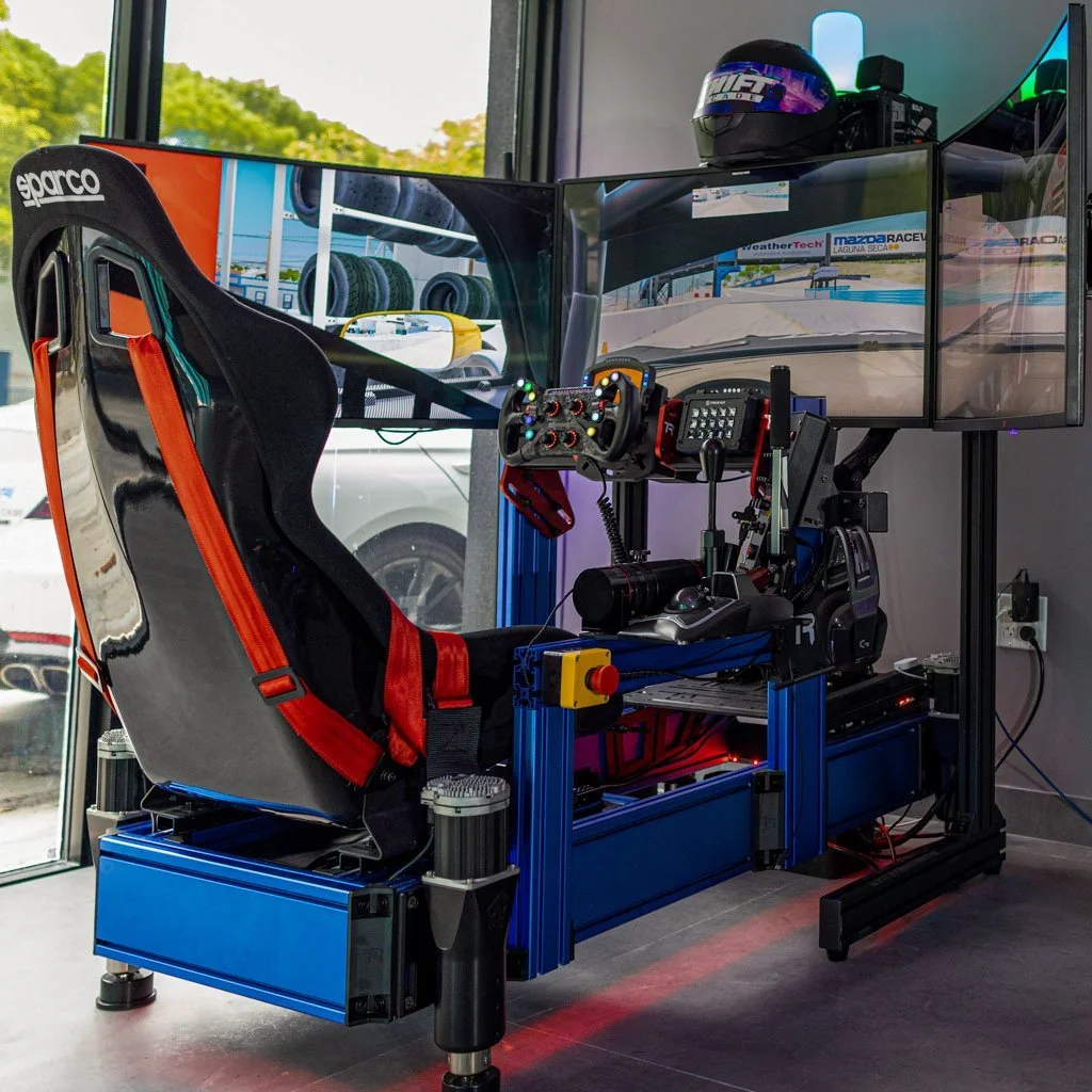 Sim racing simulator setup with a racing seat, multiple monitors displaying a race track, and various racing controls in a room with a large window and cars outside.