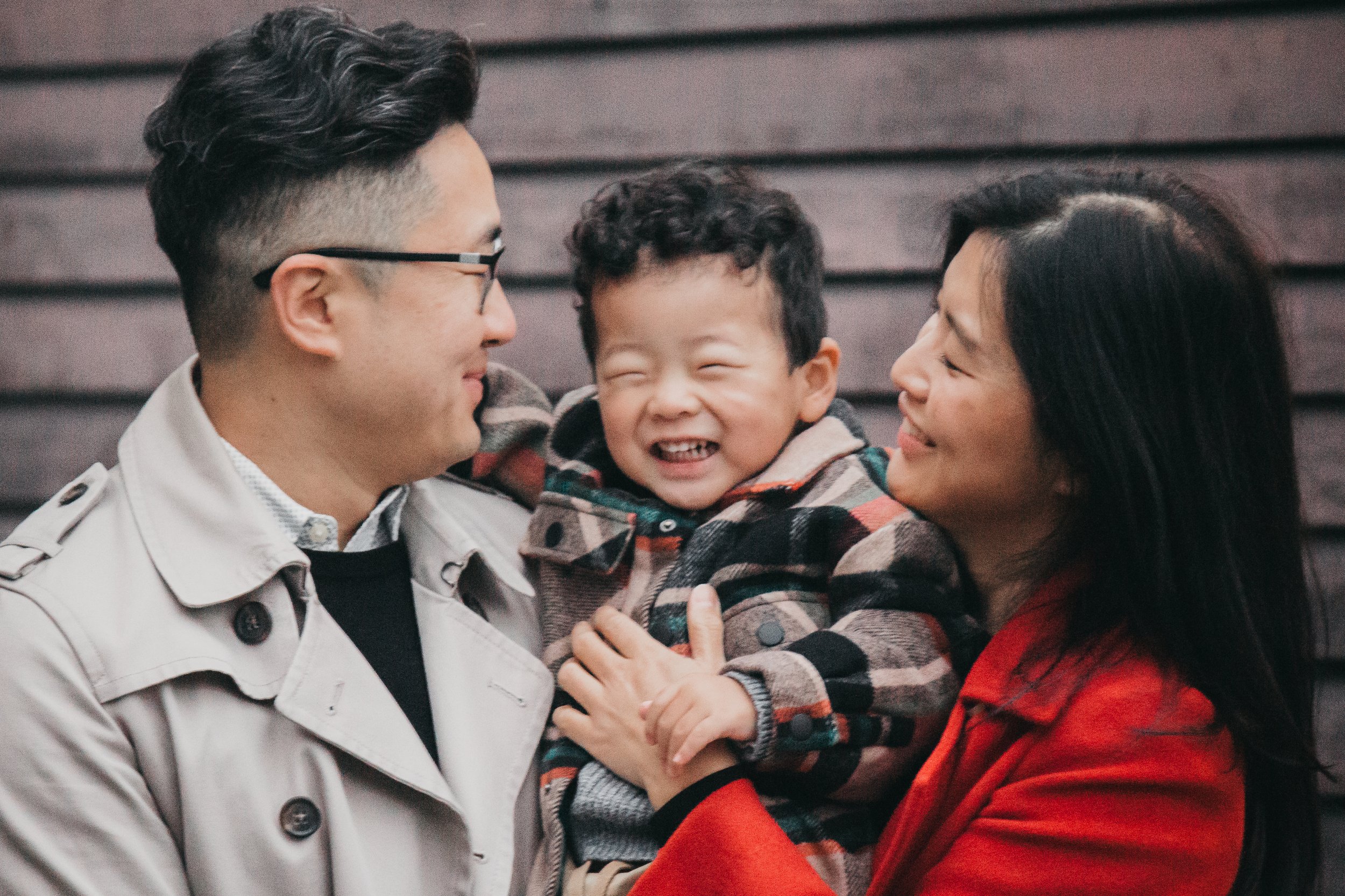 A family of three smiling, with the parents holding their child between them. The father is wearing glasses and a beige coat, the child is in a plaid jacket, and the mother is in a red coat. The background is a wooden wall.
